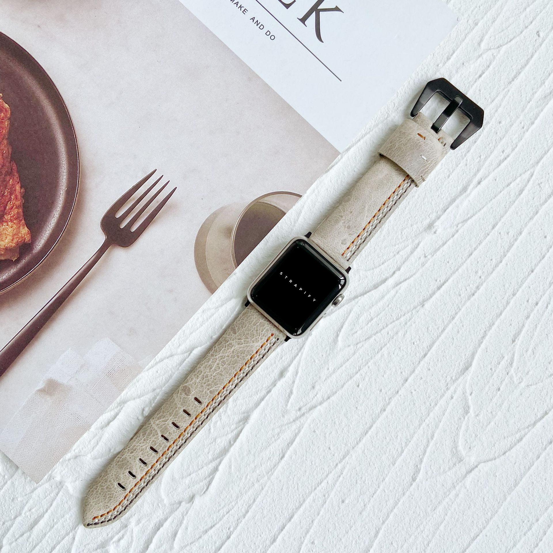 Apple Watch with ivory white leather band featuring asymmetrical stitch detail and black buckle on textured surface