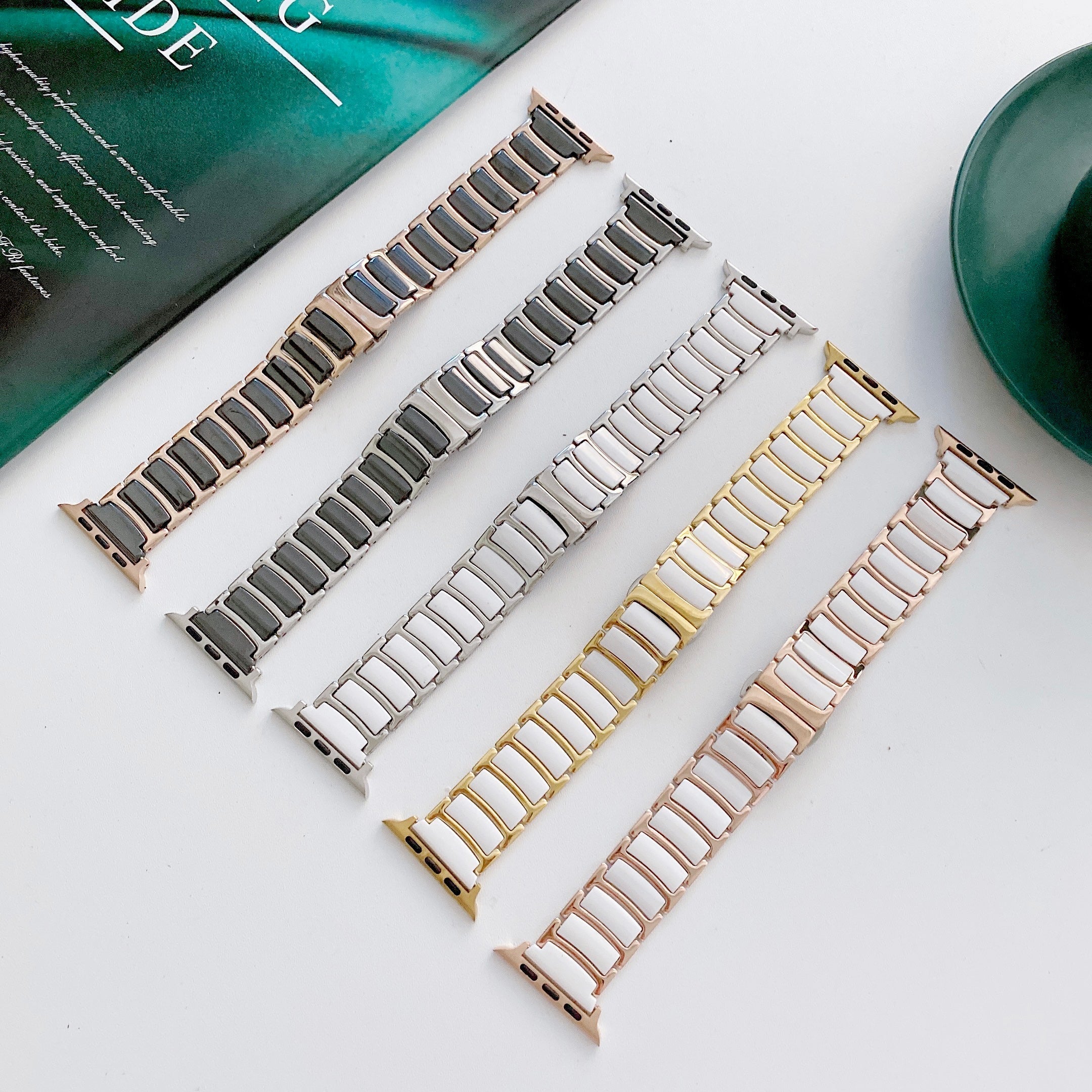 Five ceramic and steel smartwatch bands in rose gold, silver, gold, gunmetal, and blush colors displayed on white surface with green accessories