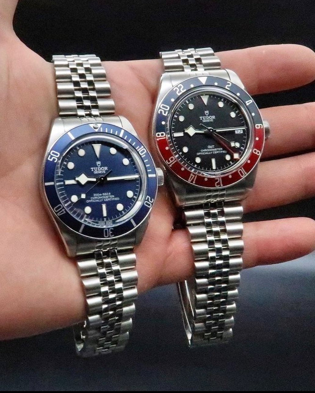 Two Tudor Black Bay stainless steel watches with blue and red bezels on jubilee bracelets displayed on wrist