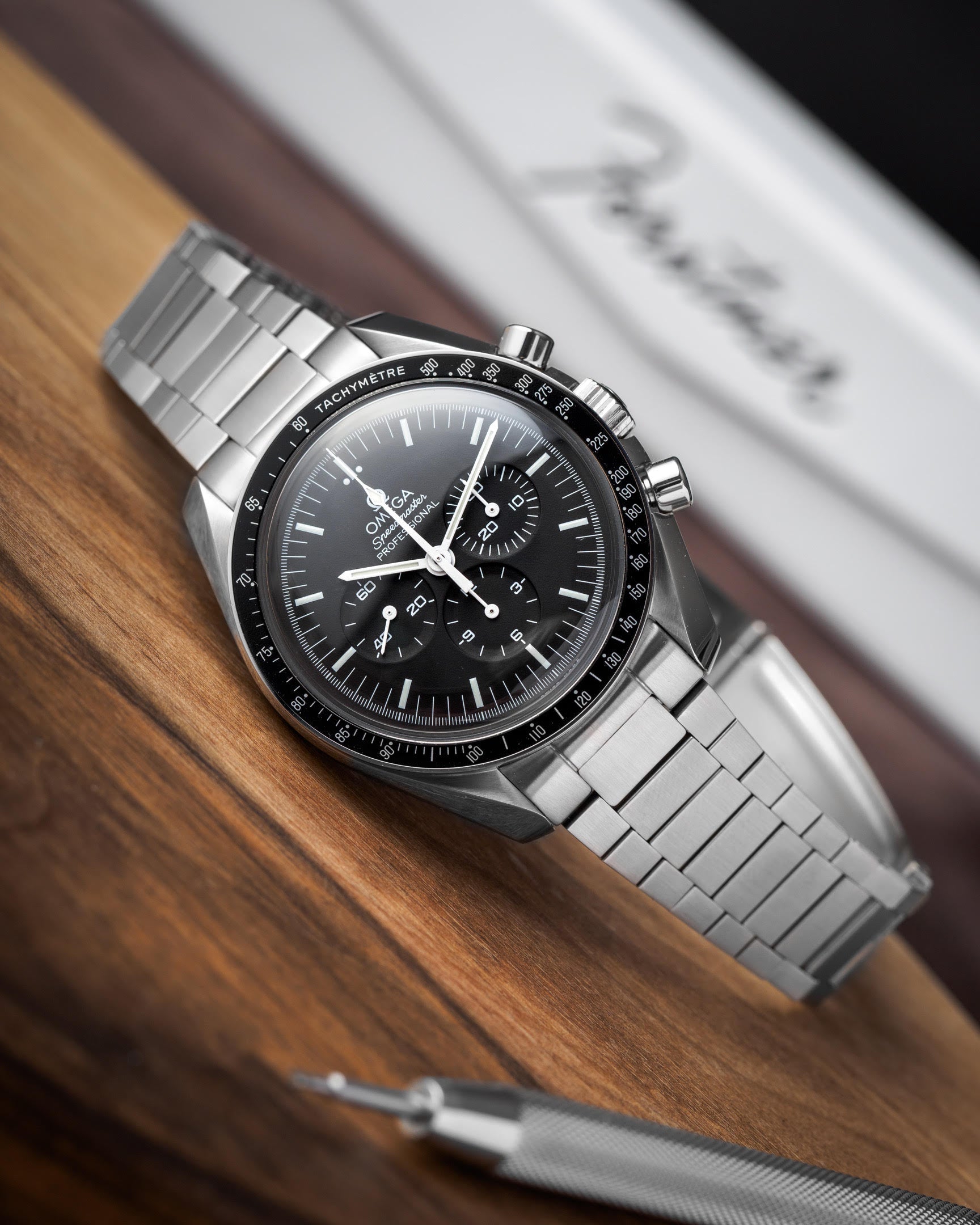 Stainless steel chronograph watch with black dial and tachymetre bezel on flat link bracelet, displayed on wooden surface