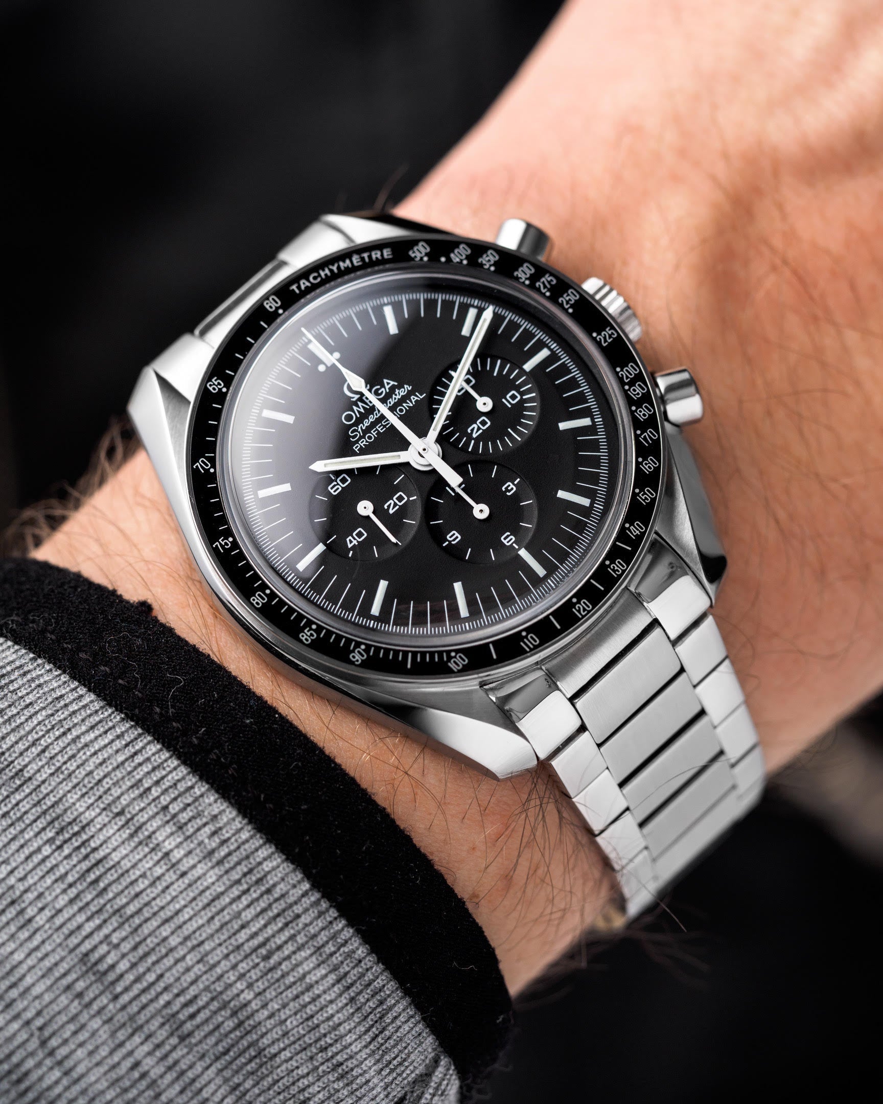 Silver stainless steel chronograph watch with black dial and tachymeter bezel on flat link bracelet worn on wrist