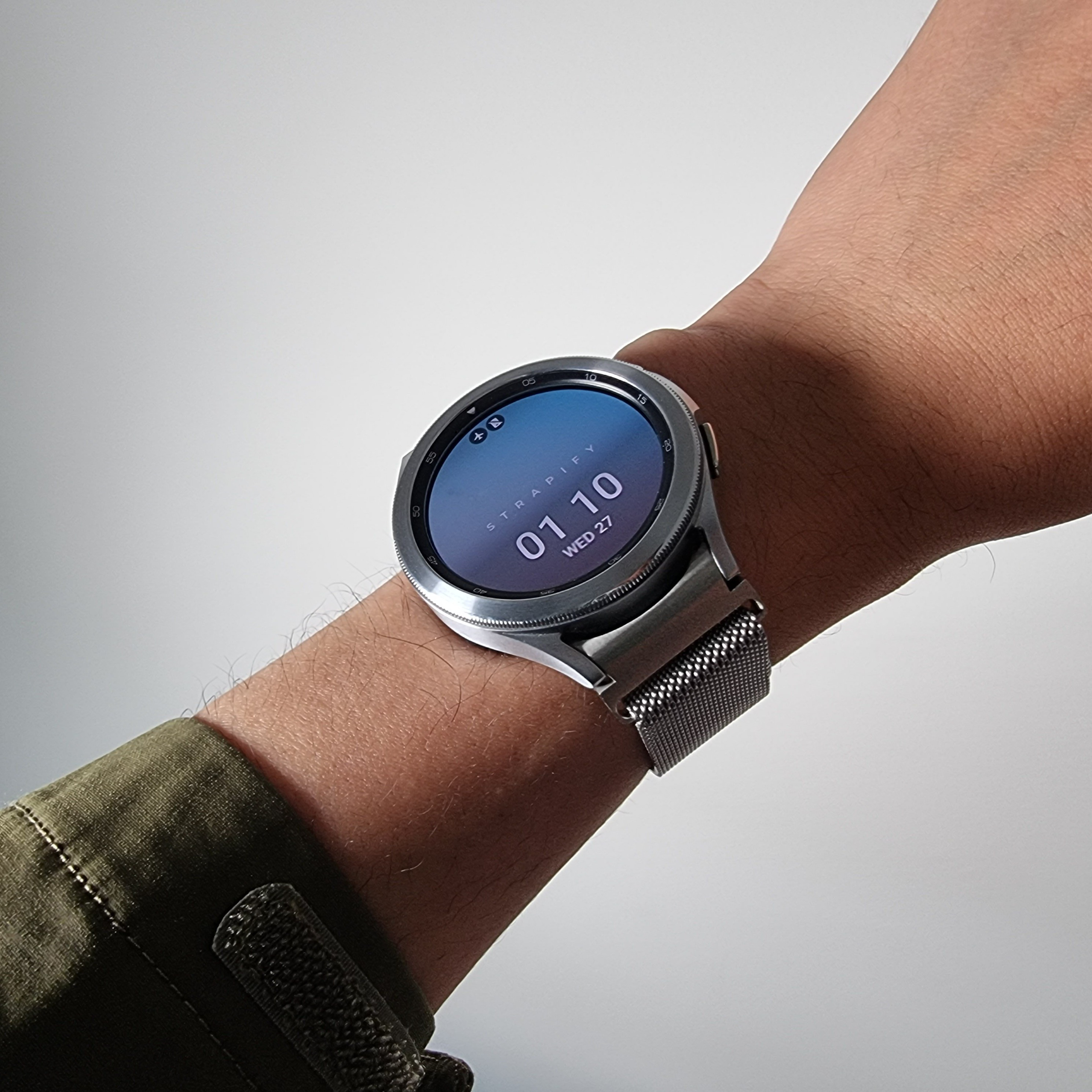 Silver Milanese mesh band smartwatch with blue digital display showing time 01:10, worn on wrist