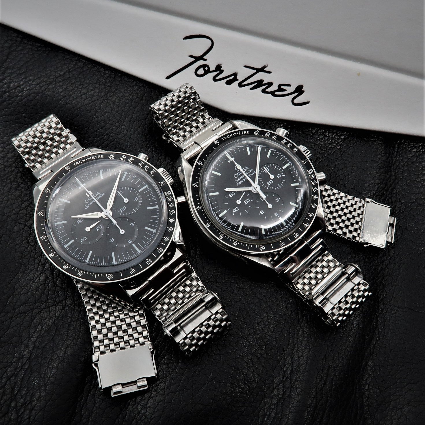 Two luxury chronograph watches with black dials and stainless steel mesh bracelets on black leather surface with Forstner branding
