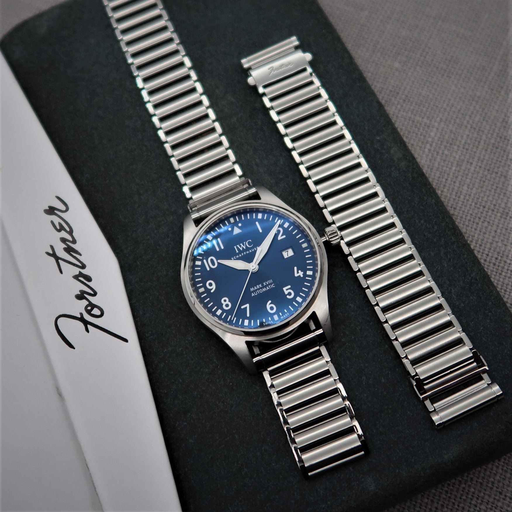 Stainless steel ladder-style watch bracelet with polished links displayed on black fabric next to IWC pilot watch with blue dial