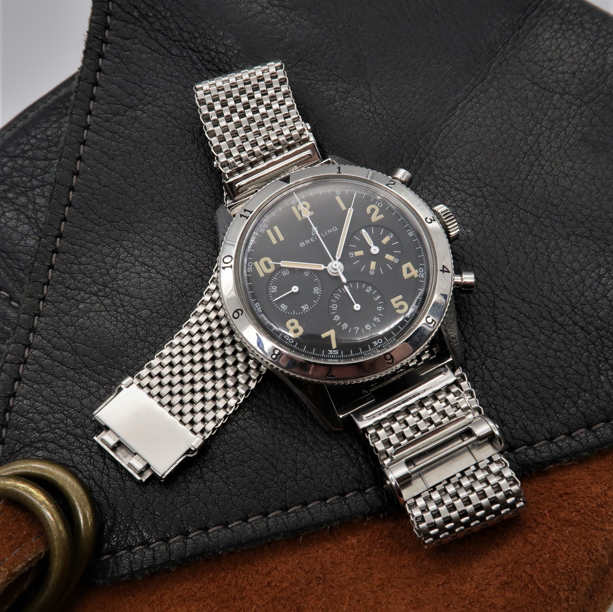 Breitling chronograph watch with black dial and Arabic numerals on stainless steel mesh bracelet displayed on black leather