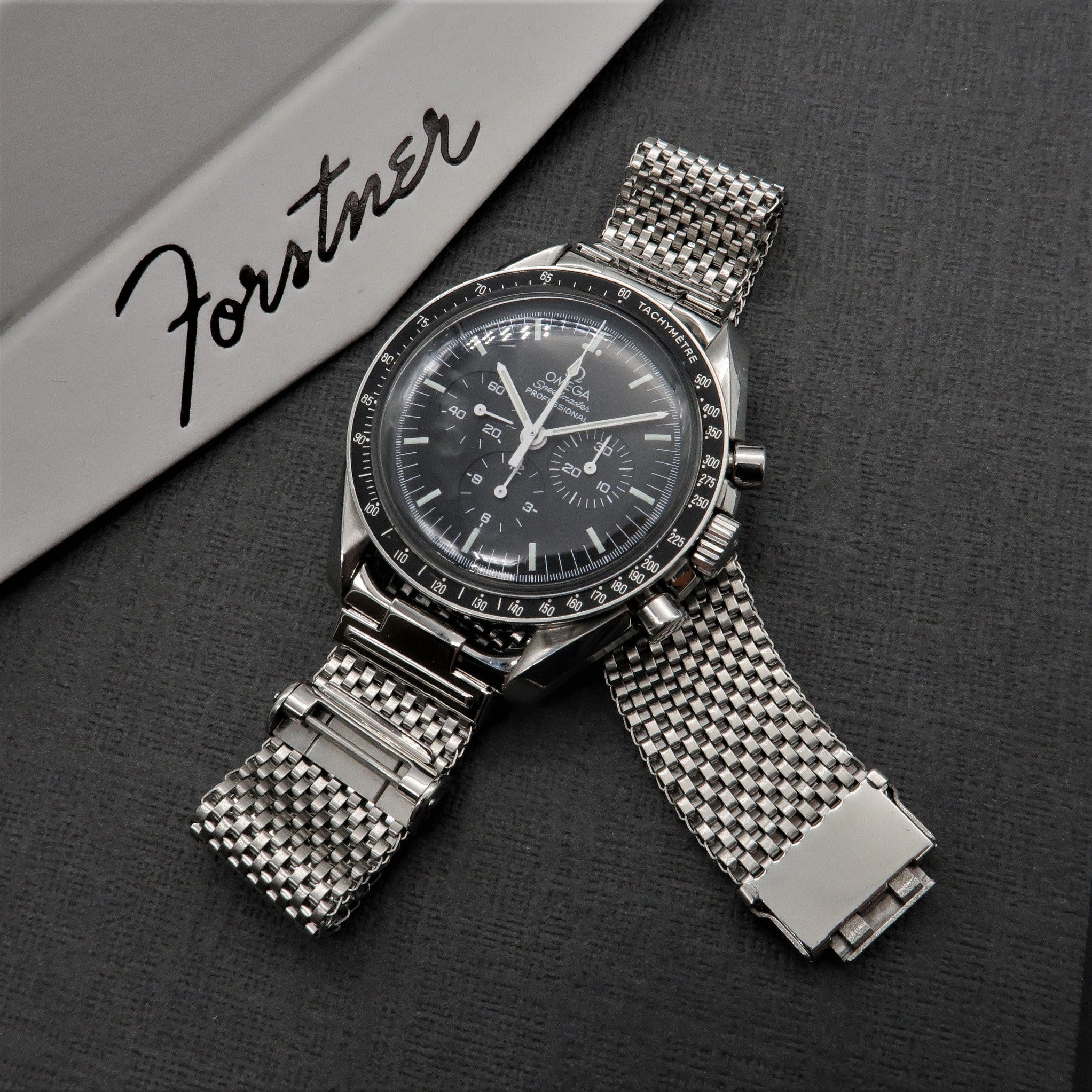 Stainless steel mesh watch bracelet with horned ends and solid end links on gray textured surface with Forster branding