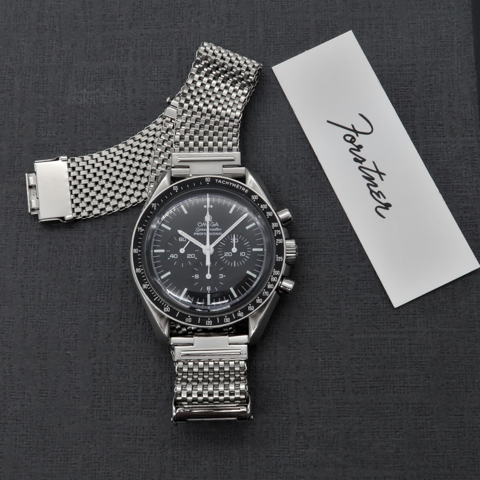 Omega Speedmaster chronograph watch with stainless steel mesh bracelet and original box on dark background