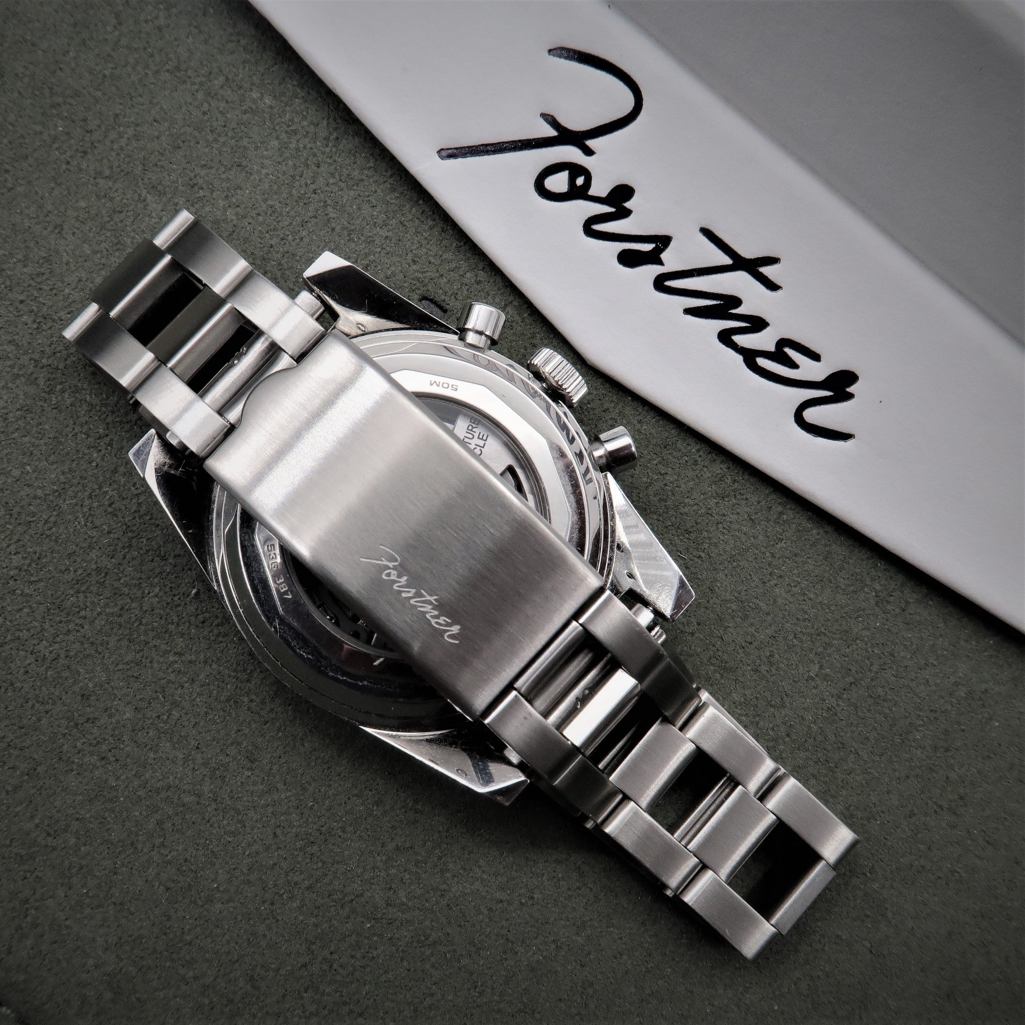Silver chronograph watch with stainless steel ladder bracelet and black dial on gray background next to product packaging