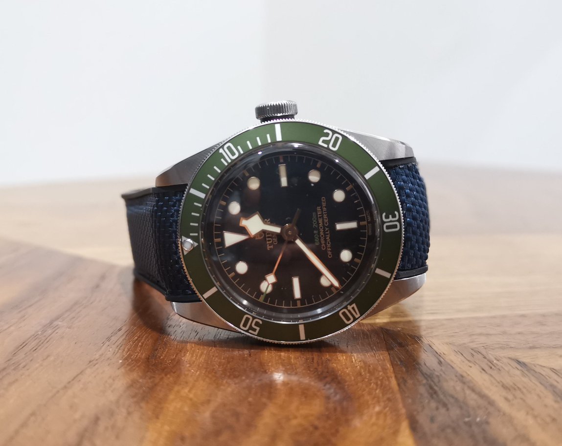 Steel dive watch with green bezel, black dial, luminous hands, and navy blue rubber strap with deployant clasp on wooden surface