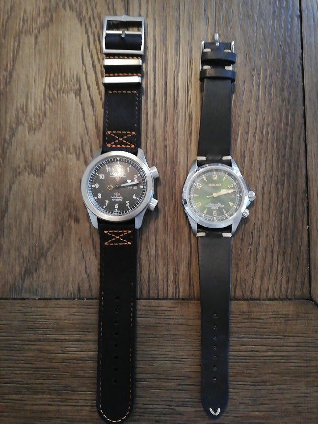 Two military-style watches with black leather straps displayed on wooden background, left watch features orange stitching
