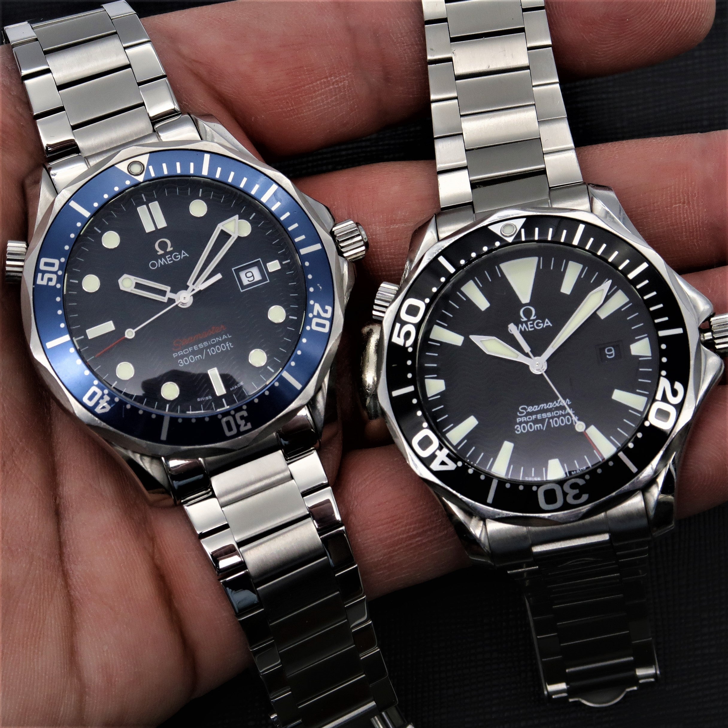 Two Omega Seamaster professional dive watches with stainless steel flat link bracelets, displayed in hand showing blue and black bezels