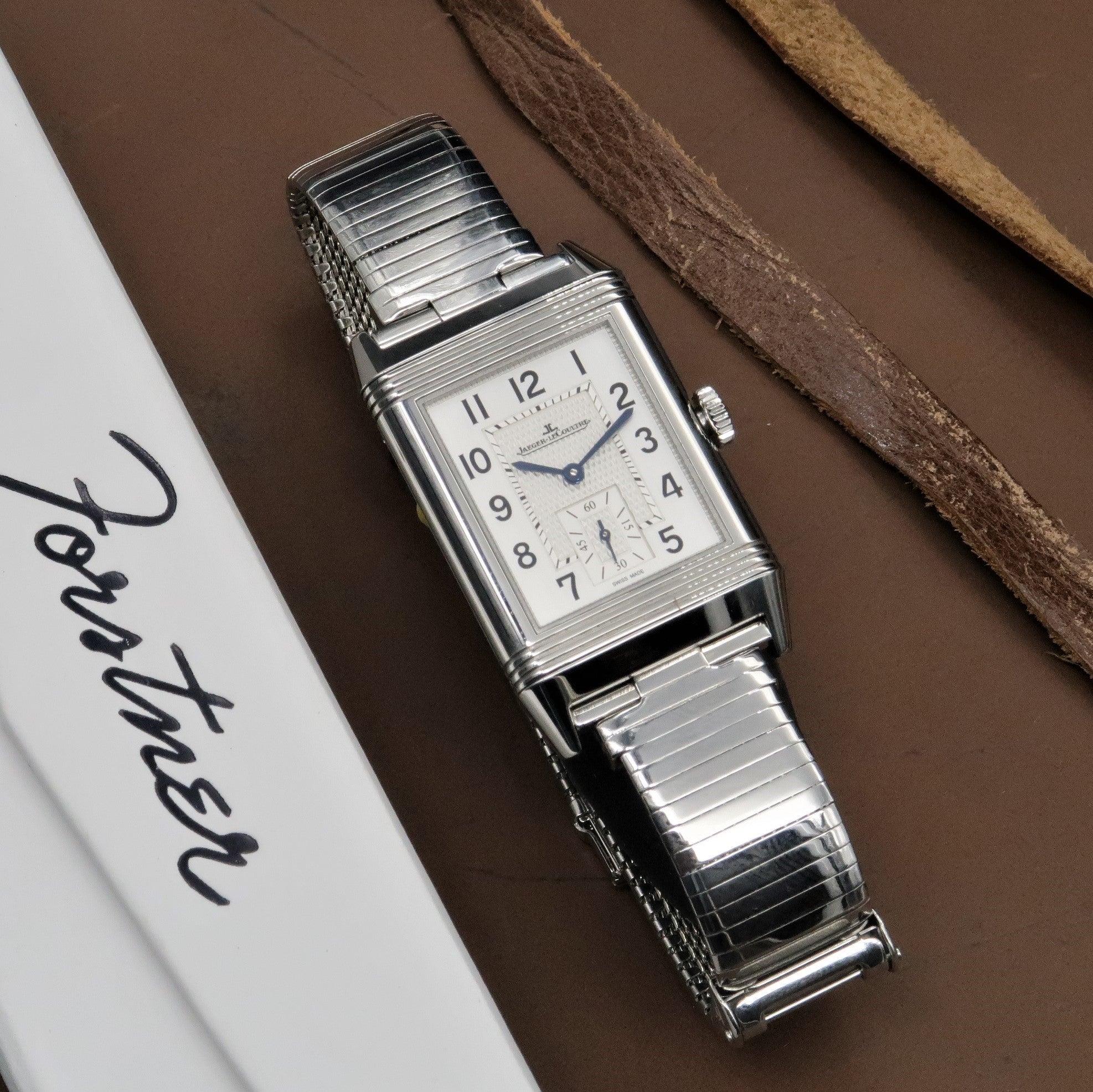 Jaeger-LeCoultre Reverso rectangular stainless steel watch with cream dial, Arabic numerals, and expandable metal bracelet on brown background