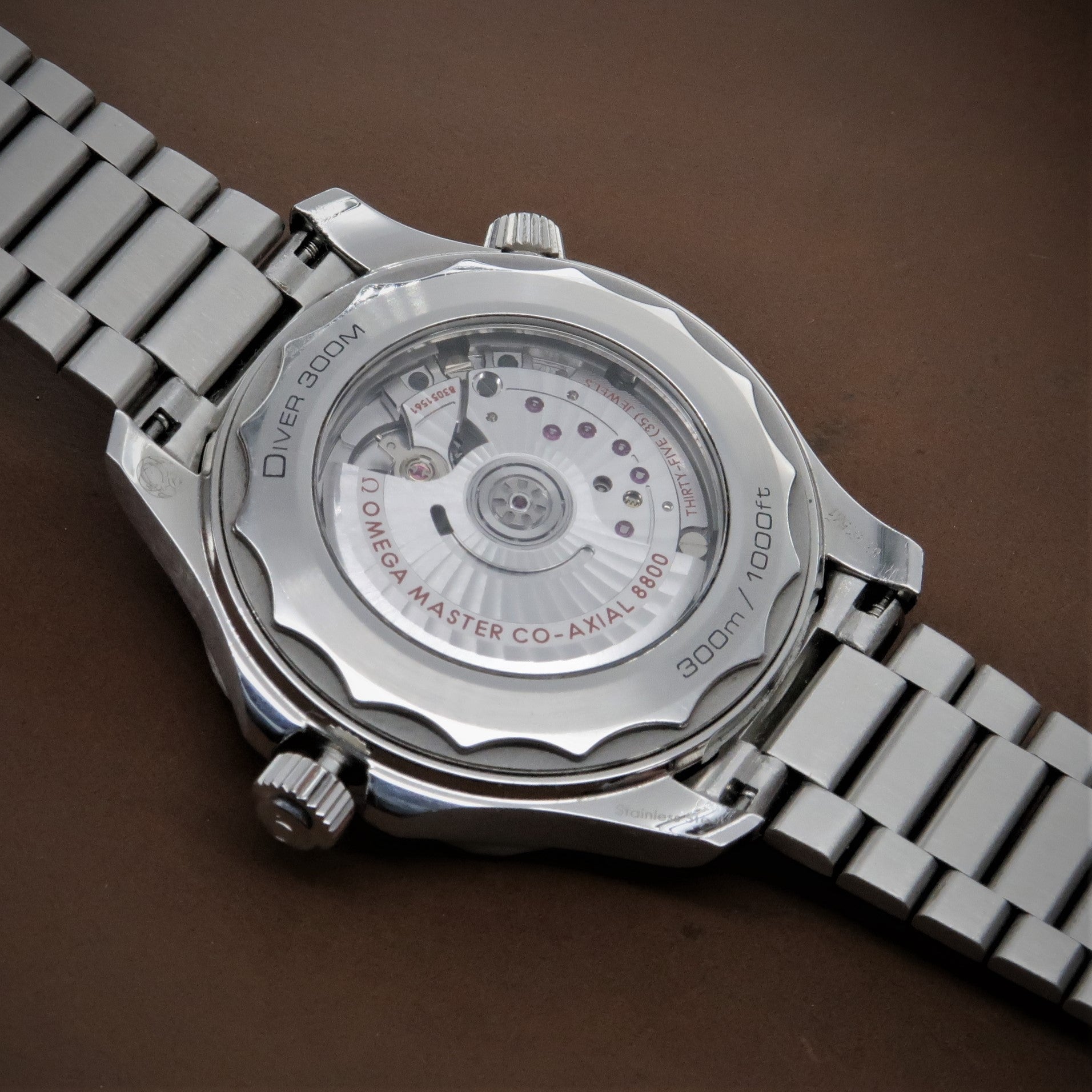 Silver stainless steel dive watch with exposed caseback showing co-axial movement, metal bracelet, water resistant to 300m