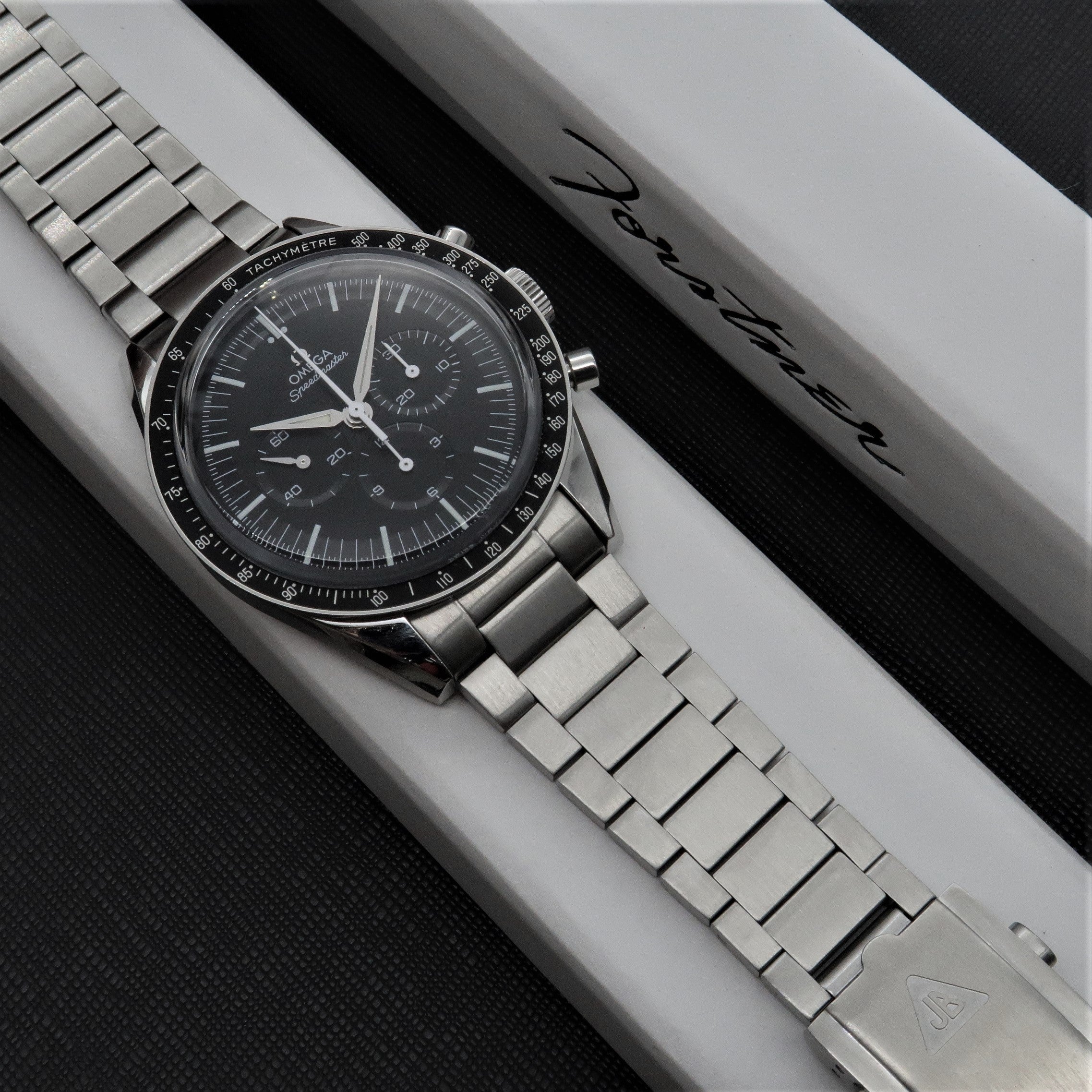 Omega Speedmaster chronograph watch with black dial and stainless steel Forstner flat link bracelet on white display box