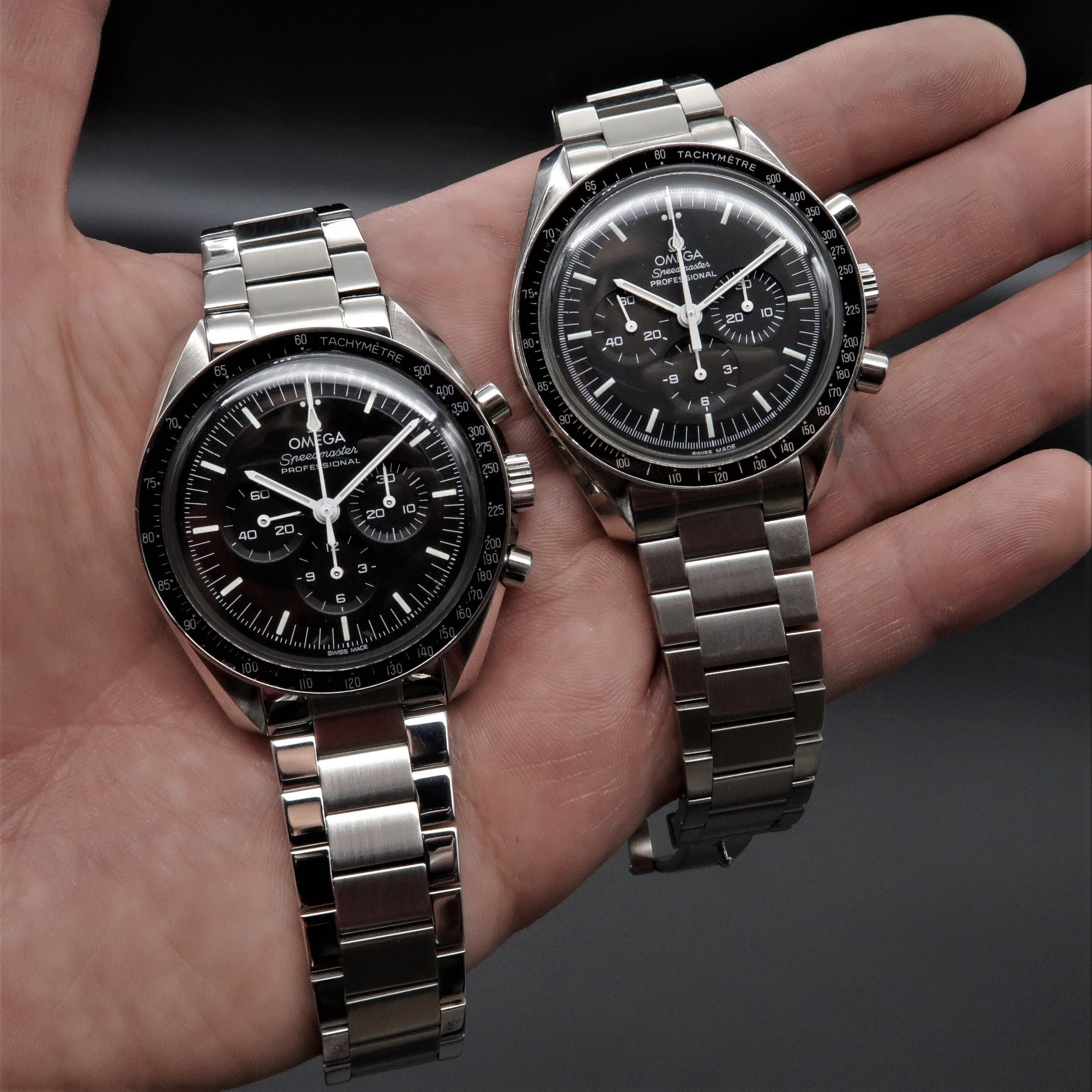 Two Omega Speedmaster chronograph watches with stainless steel flat link bracelets displayed in hand, featuring black dials and tachymeter bezels