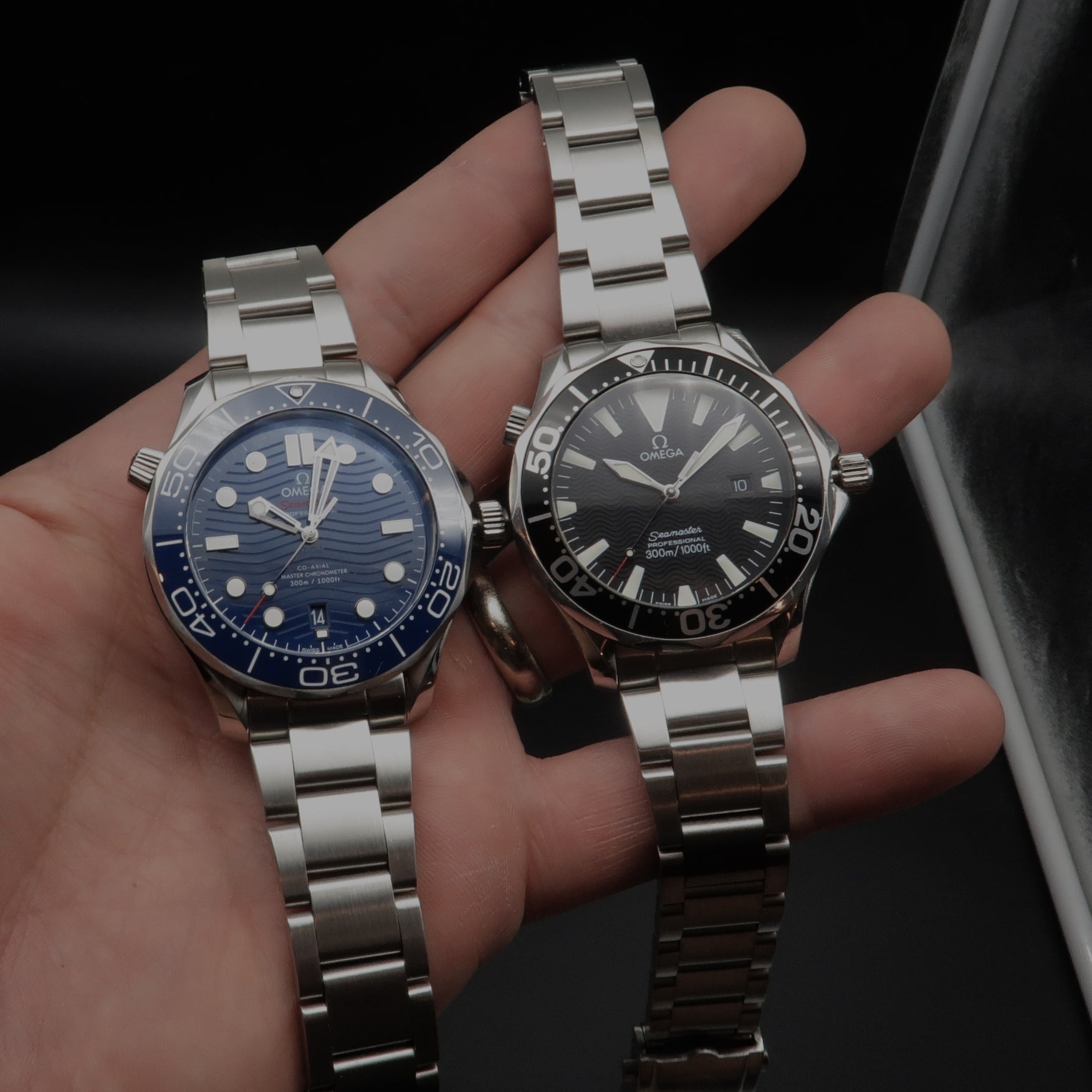 Two Omega Seamaster dive watches with stainless steel bracelets, one with blue dial and one with black dial, displayed in hand