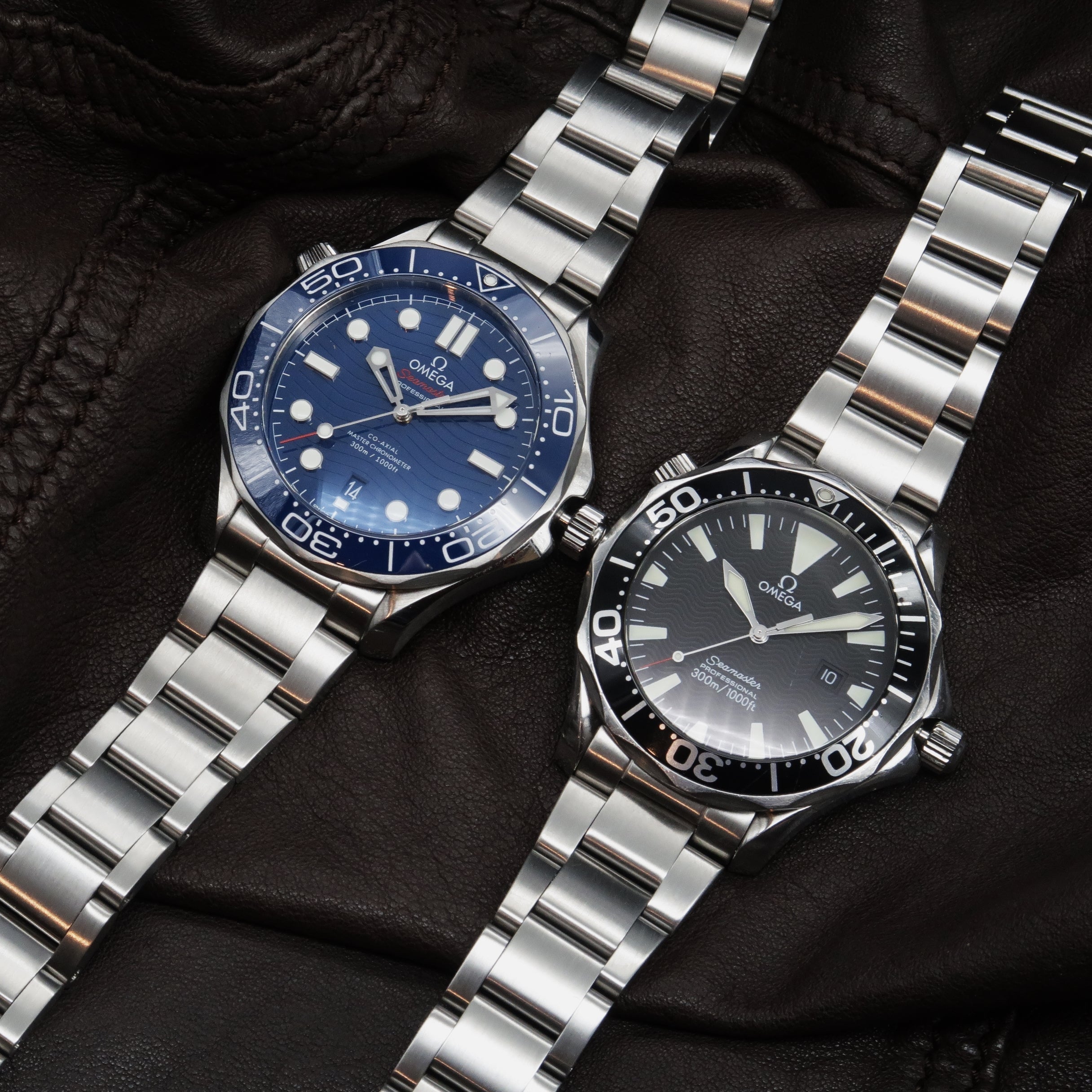 Two Omega Seamaster dive watches with stainless steel bracelets, one with blue dial and one with black dial, displayed on black leather background