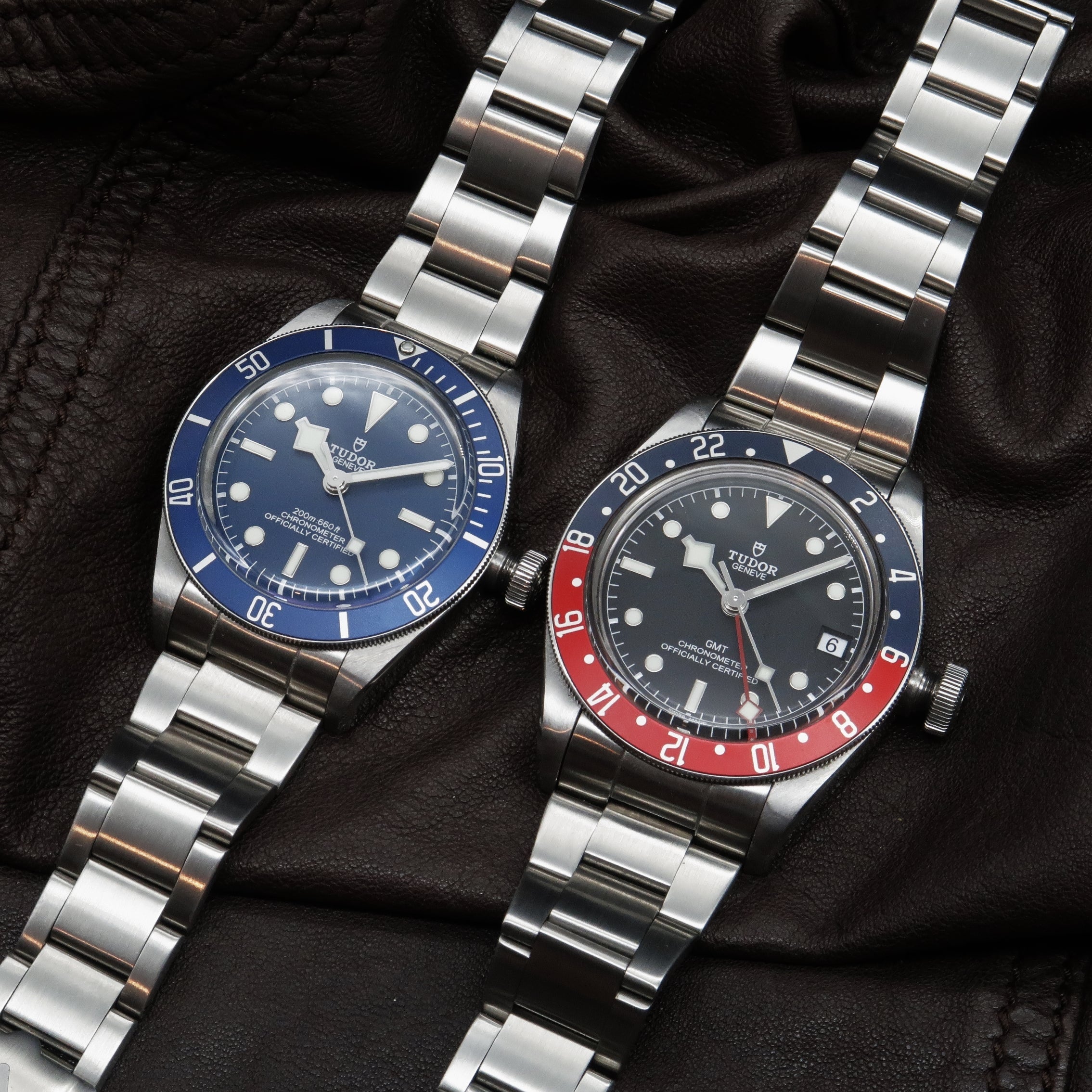 Two Tudor Black Bay steel dive watches with blue and red GMT bezels on matching bracelets against black fabric
