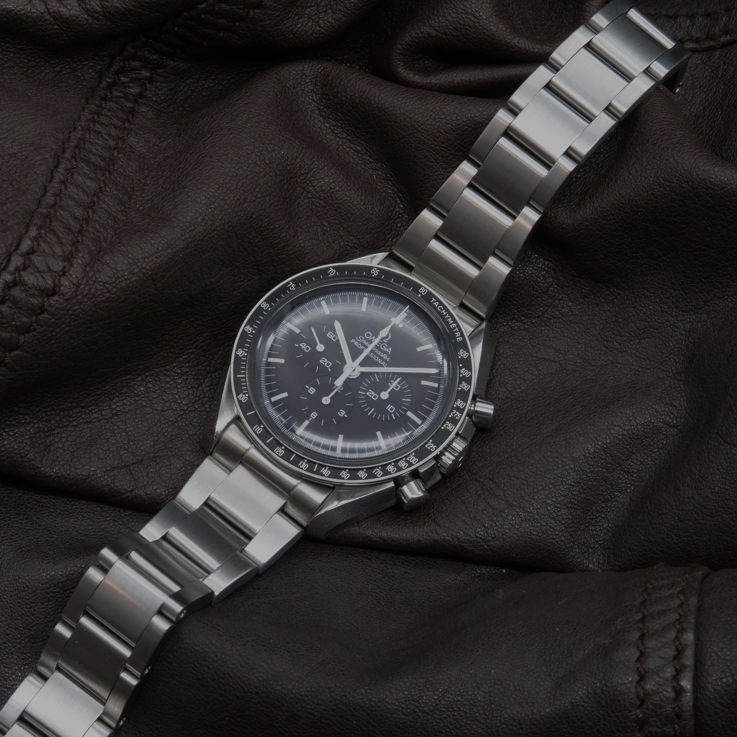 Stainless steel chronograph watch with black dial, tachymeter bezel, and metal bracelet on black leather background