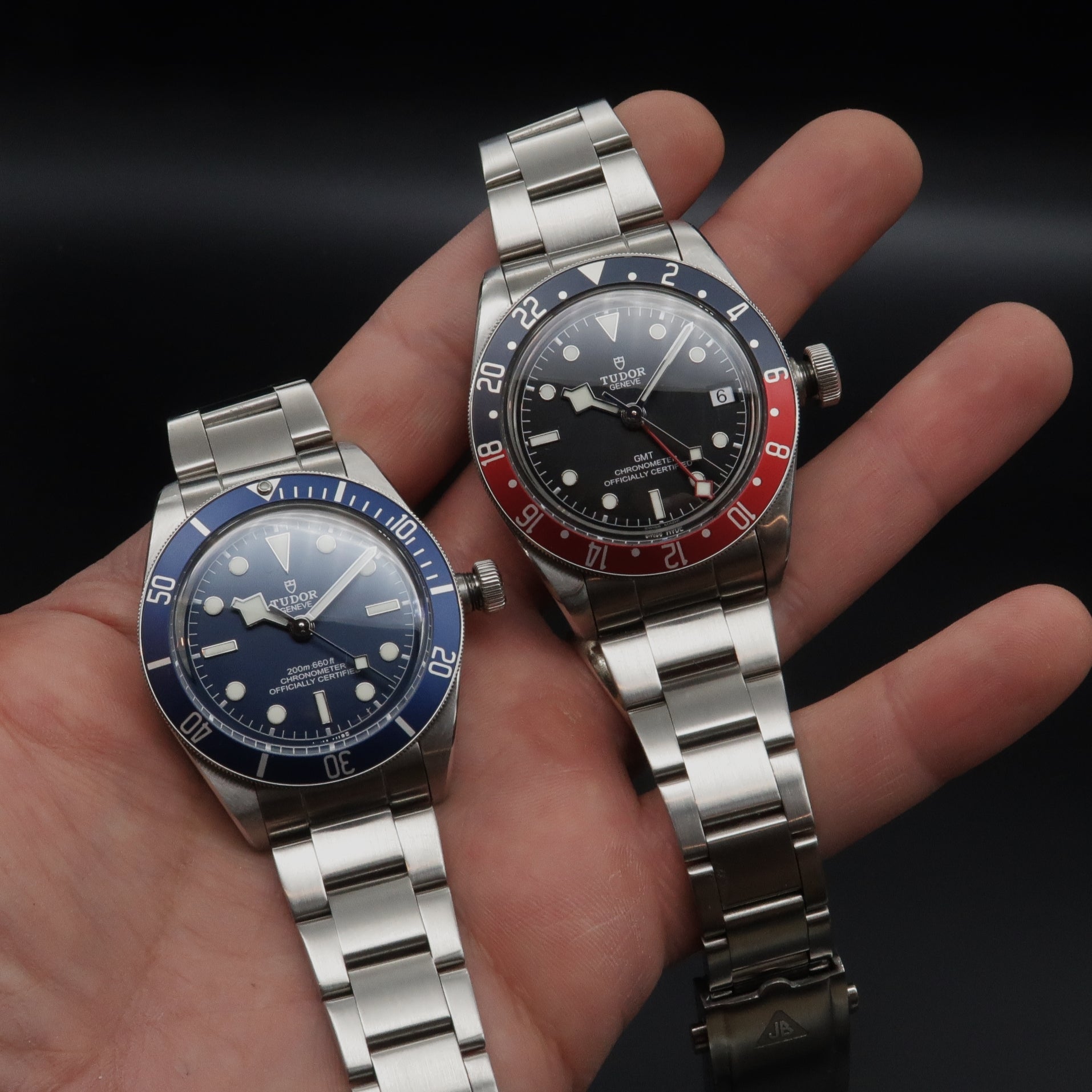 Two Tudor Black Bay luxury watches with stainless steel bracelets, one with blue bezel and black dial, one with red-blue GMT bezel and black dial, held in hand