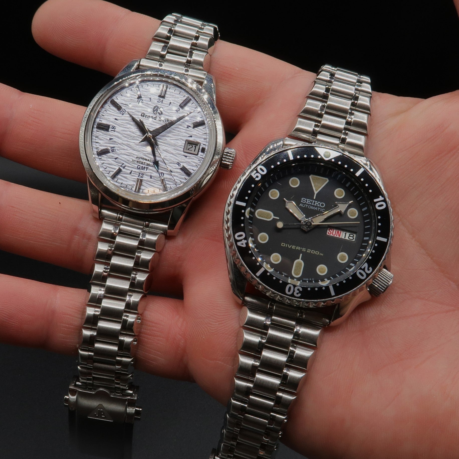 Pair of luxury automatic watches with steel bracelets held in hand: white dial Grand Seiko GMT and black dial Seiko diver's watch