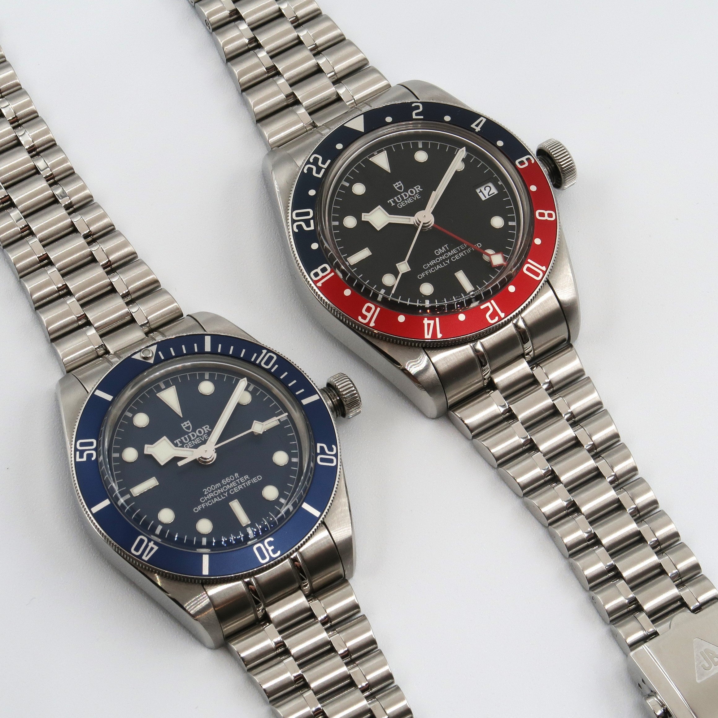 Two Tudor Black Bay dive watches with steel bracelets, one with blue bezel and one with red-black GMT bezel on white background