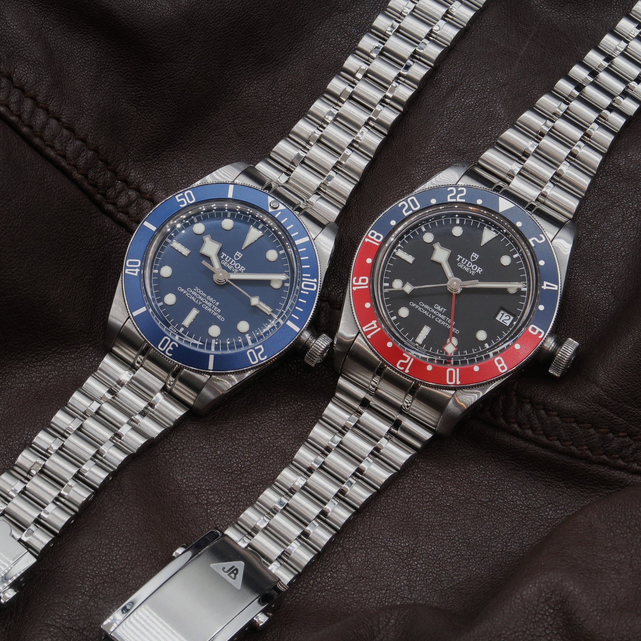 Two Tudor Black Bay steel sports watches with blue and red dive bezels on stainless steel bracelets displayed on black leather background