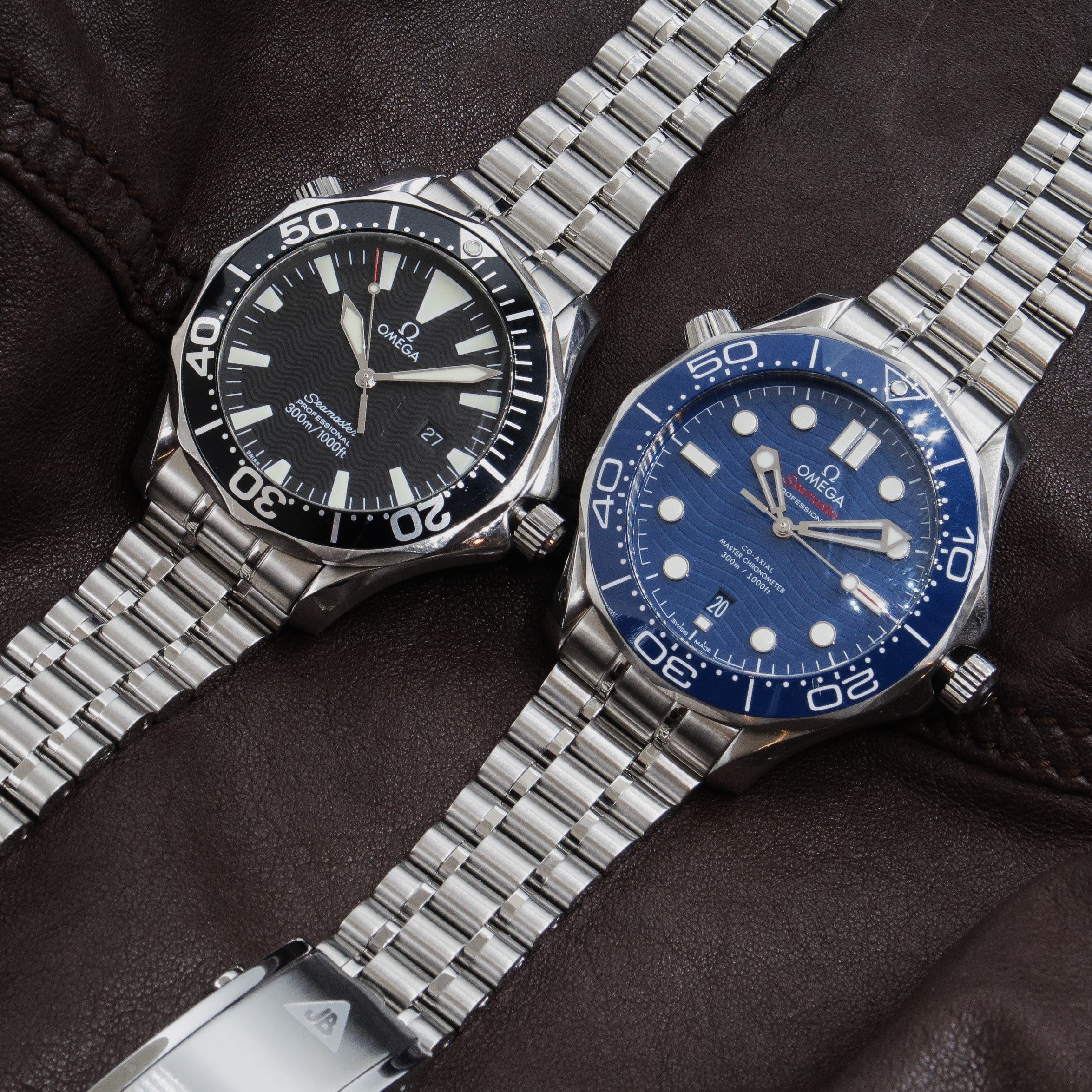 Two stainless steel Omega Seamaster Professional 300M dive watches with metal bracelets on dark leather background, black and blue dial versions