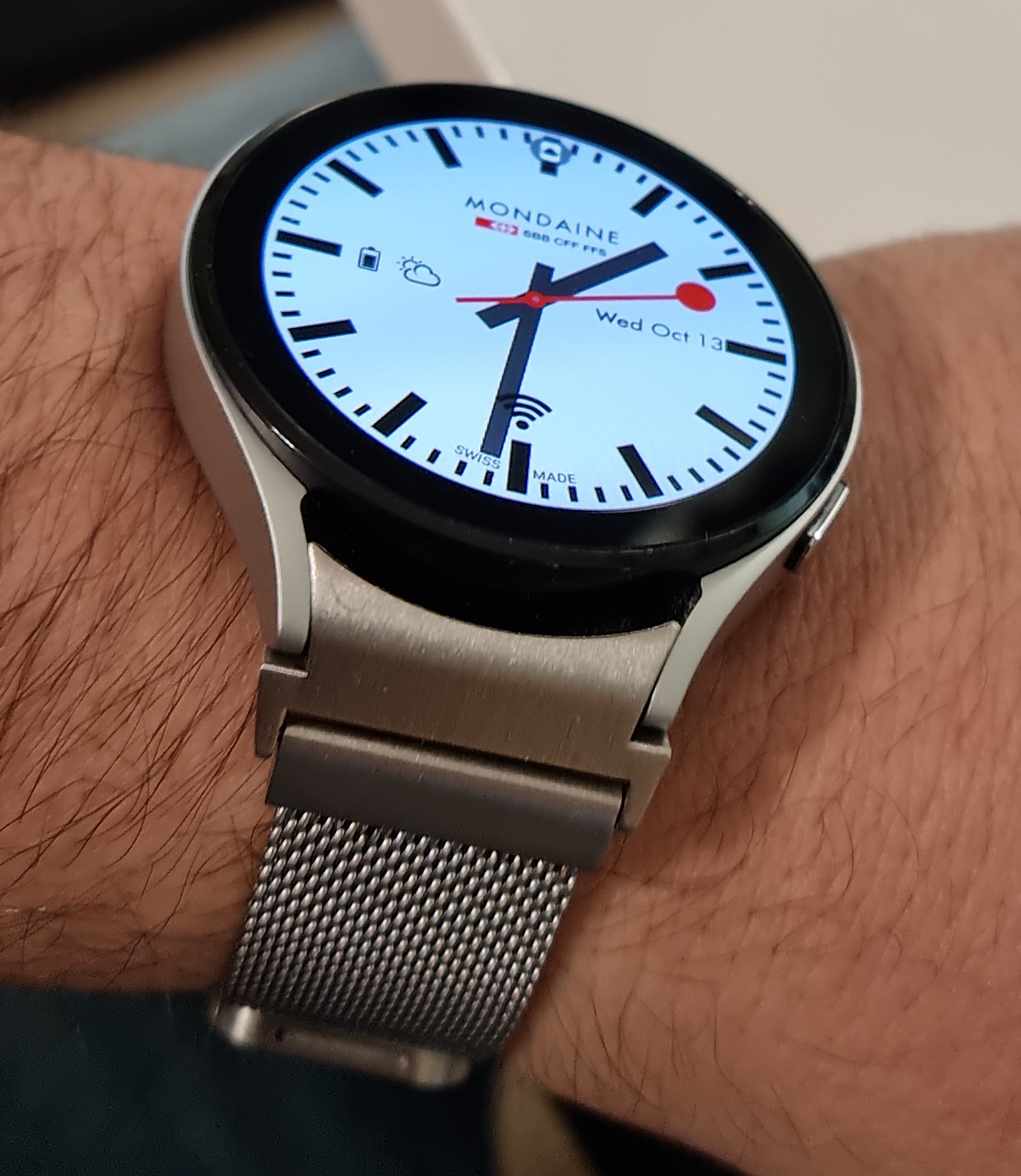 Mondaine smartwatch with stainless steel mesh band worn on wrist, light blue dial with date display and red accents
