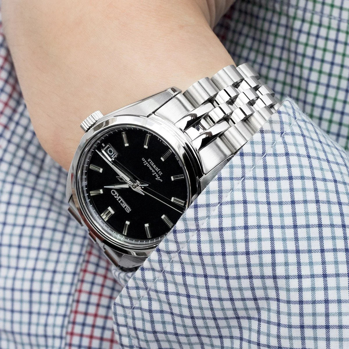 Stainless steel jubilee bracelet watch with black dial and date window on wrist wearing checkered shirt