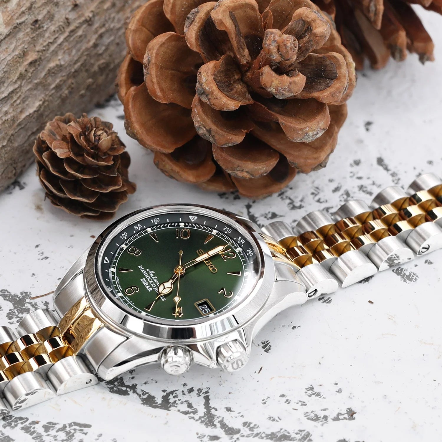 Two-tone stainless steel and gold watch bracelet with green dial watch displayed with pine cones on textured white surface