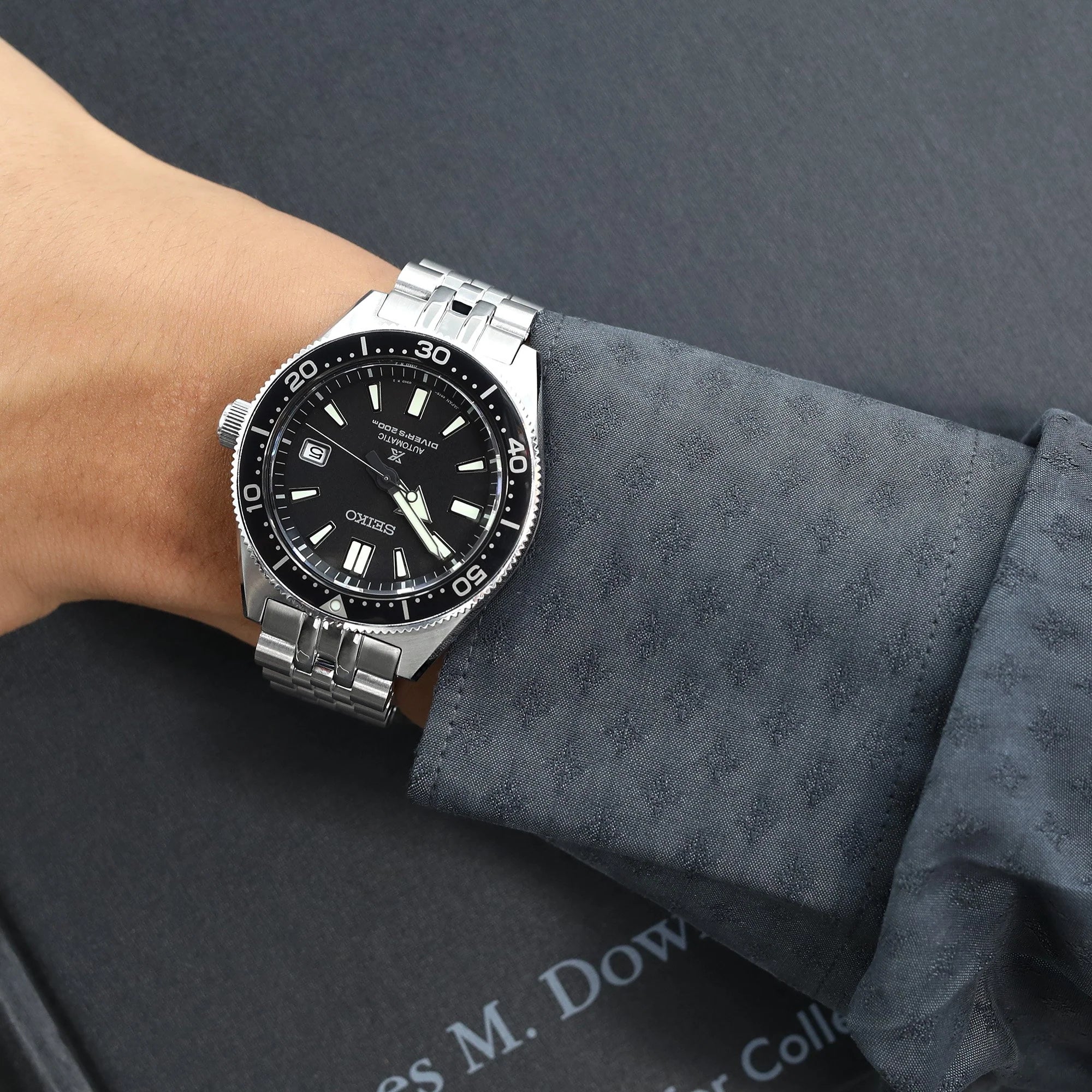Seiko dive watch with black dial and date window on stainless steel bracelet worn on wrist with gray fabric sleeve