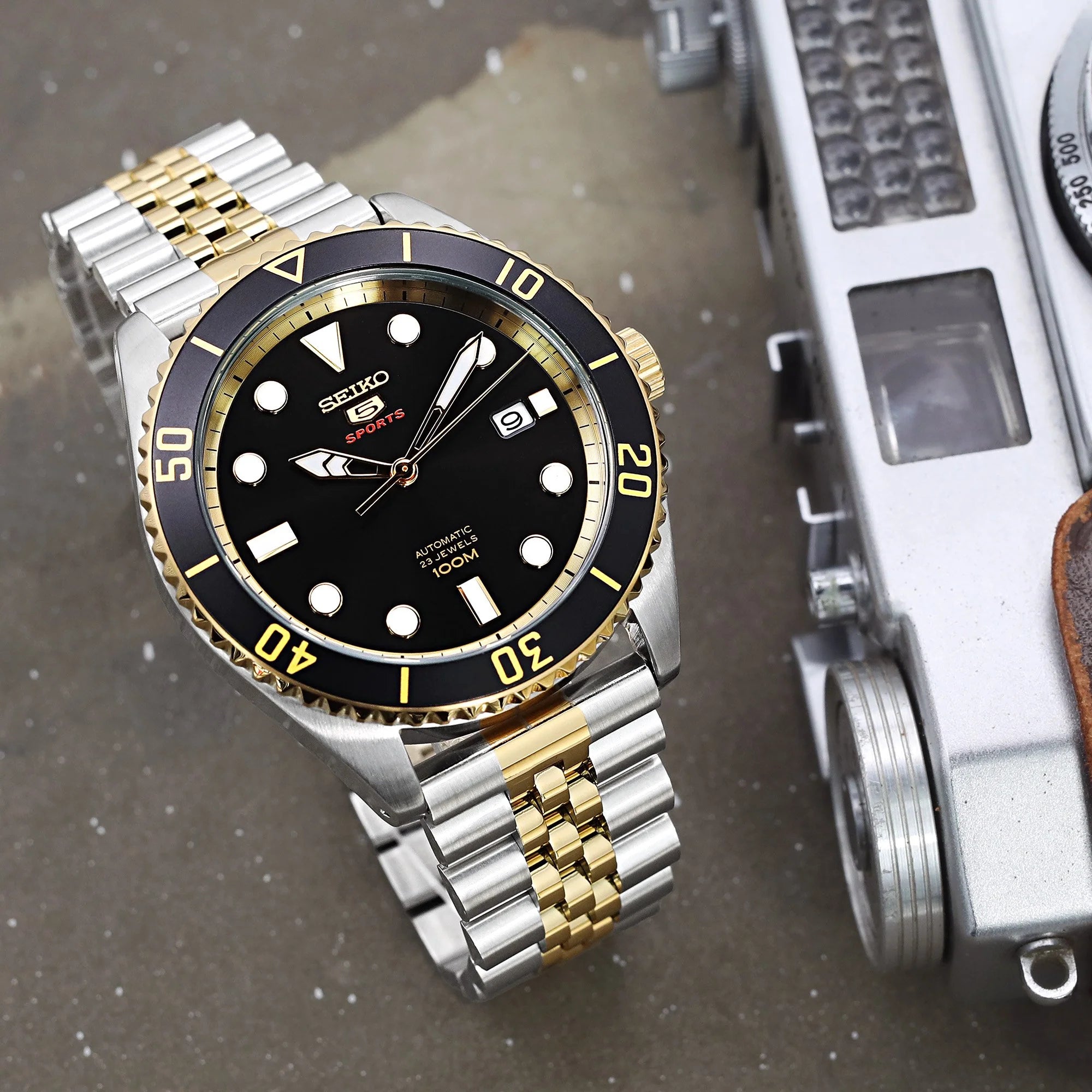 Seiko 5 Sports automatic watch with two-tone silver and gold bracelet, black dial, and rotating bezel on measurement scale background