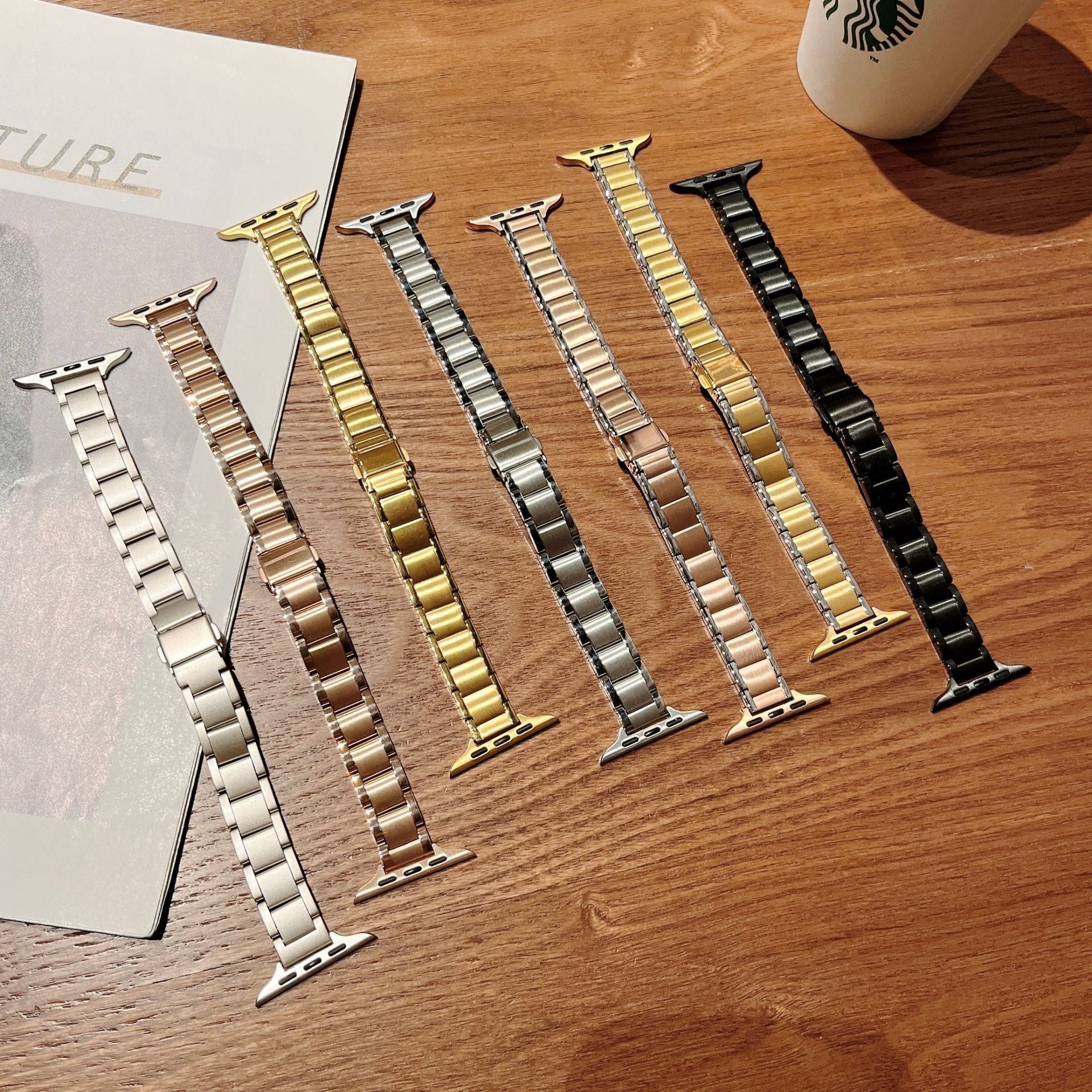 Collection of seven metal Apple Watch bands in silver, gold, rose gold, champagne, and black finishes displayed on wood surface