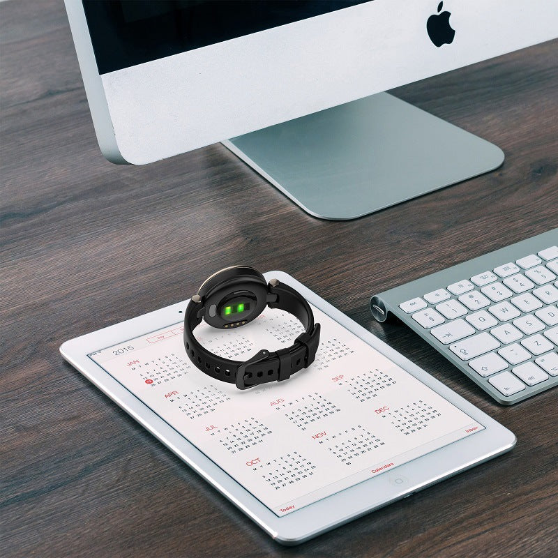 Black silicone smartwatch with green display face on calendar, beside white keyboard and iMac on wooden desk workspace