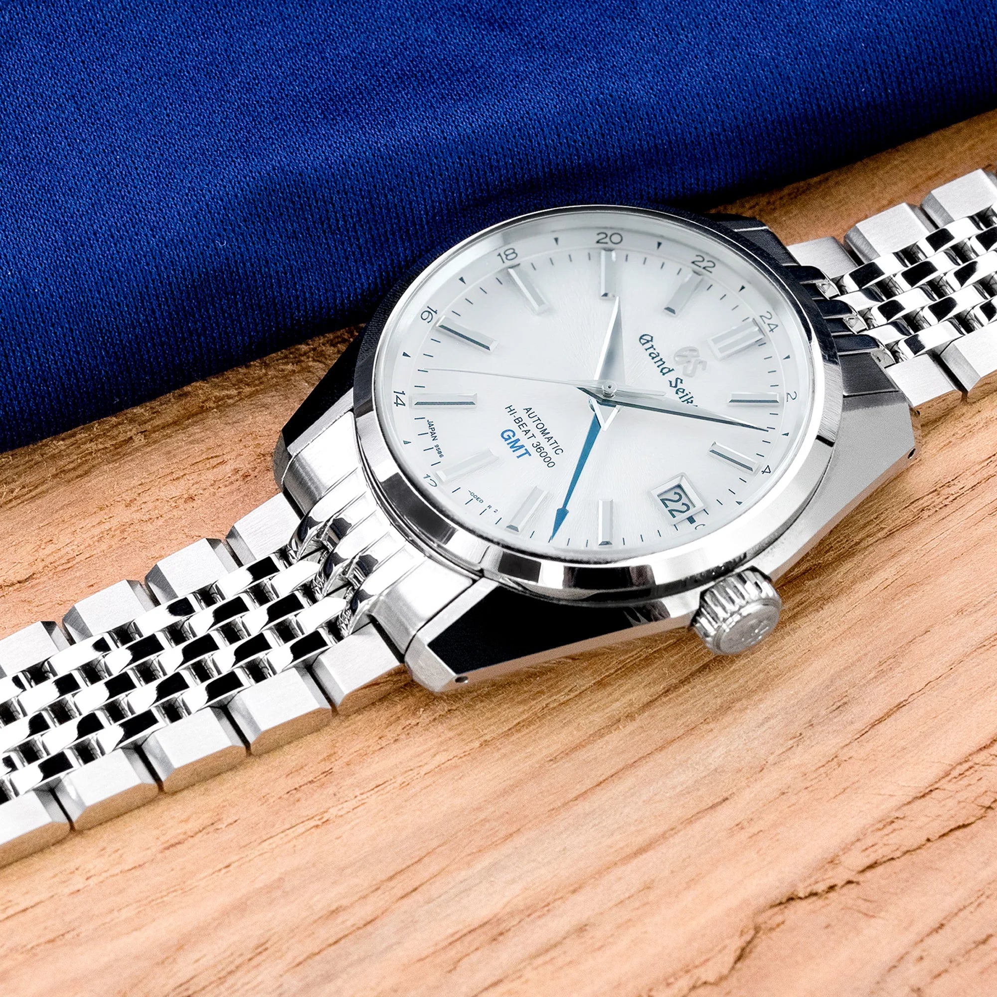 Stainless steel watch with white dial and blue accents on metal bracelet, displayed on wooden surface with blue fabric