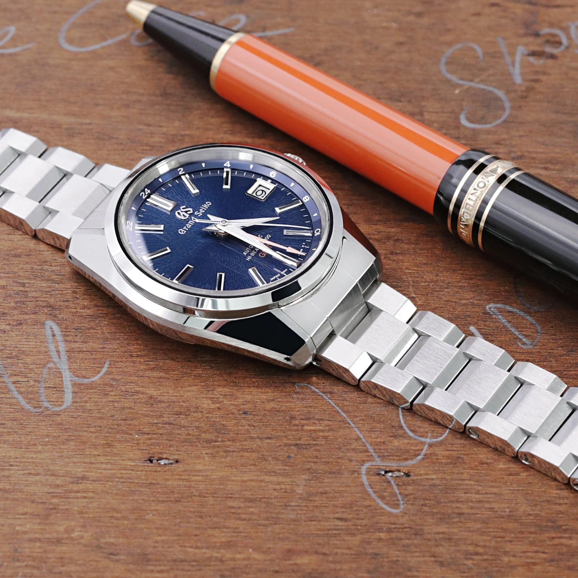 Silver stainless steel Hexad III bracelet for Grand Seiko 44GS watch displayed with orange pen on wooden desk