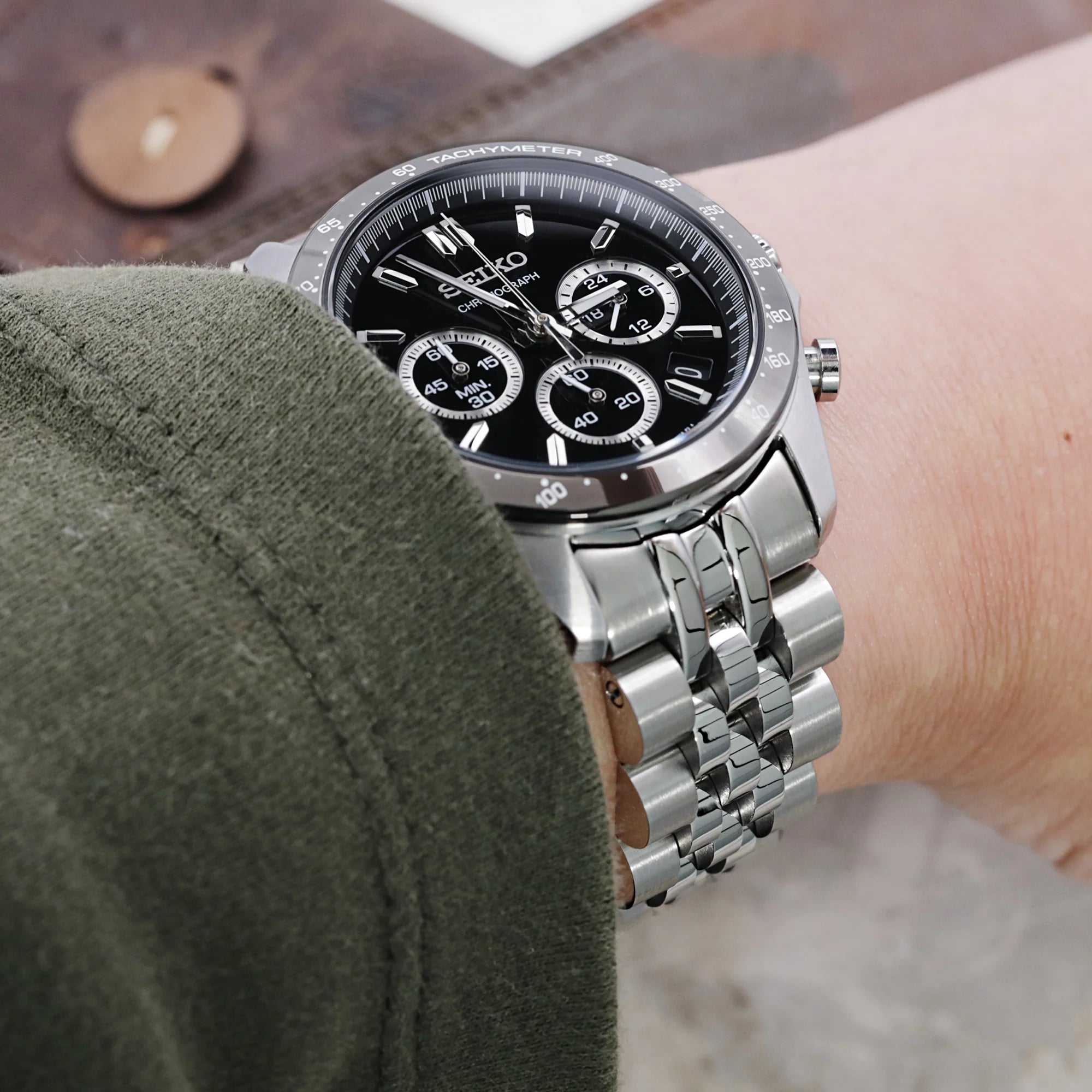 Seiko chronograph watch with black dial and three subdials on stainless steel bracelet worn on wrist with green sleeve