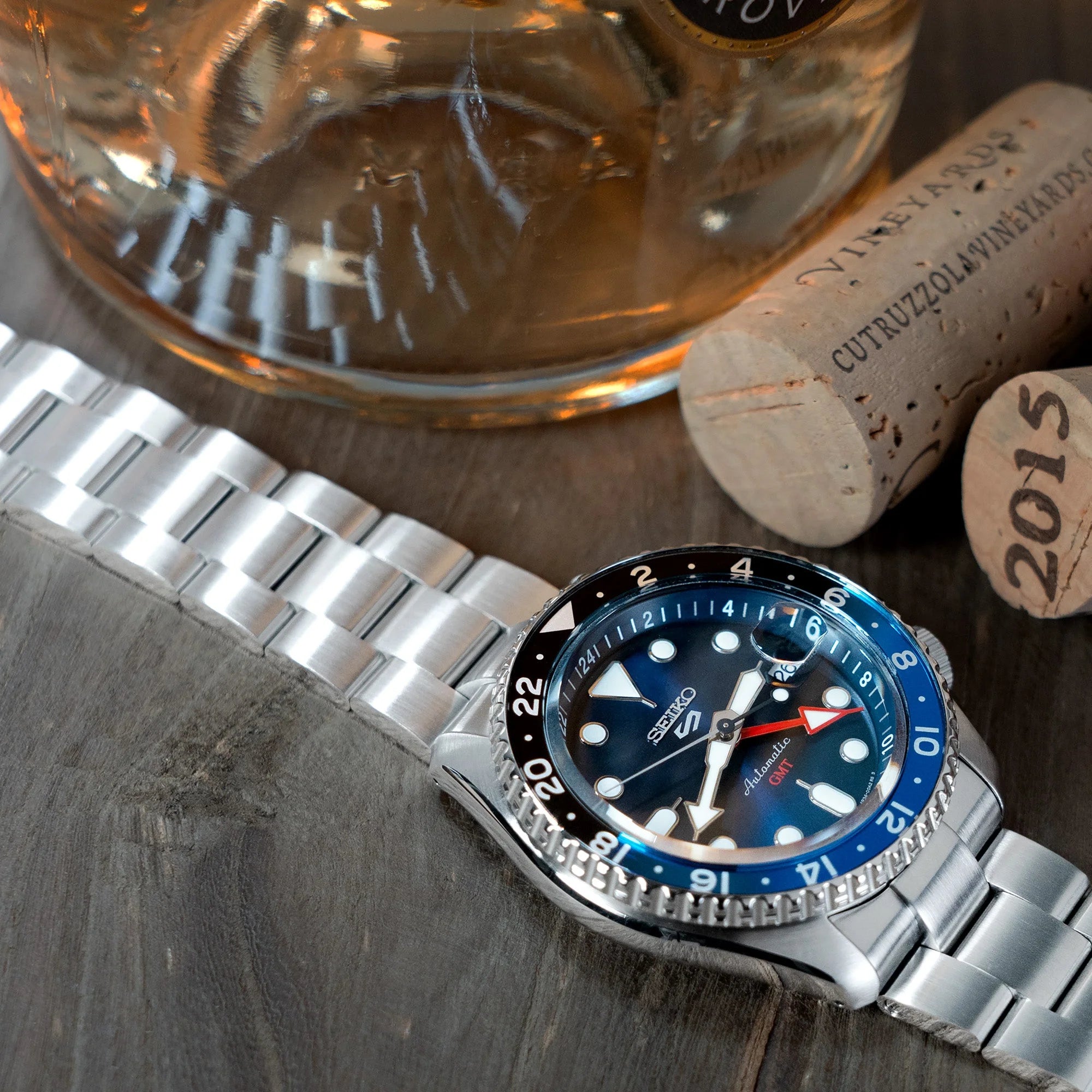 Seiko 5 GMT SSK001 dive watch with stainless steel Super-O Boyer bracelet, blue bezel, next to cognac bottle and cork