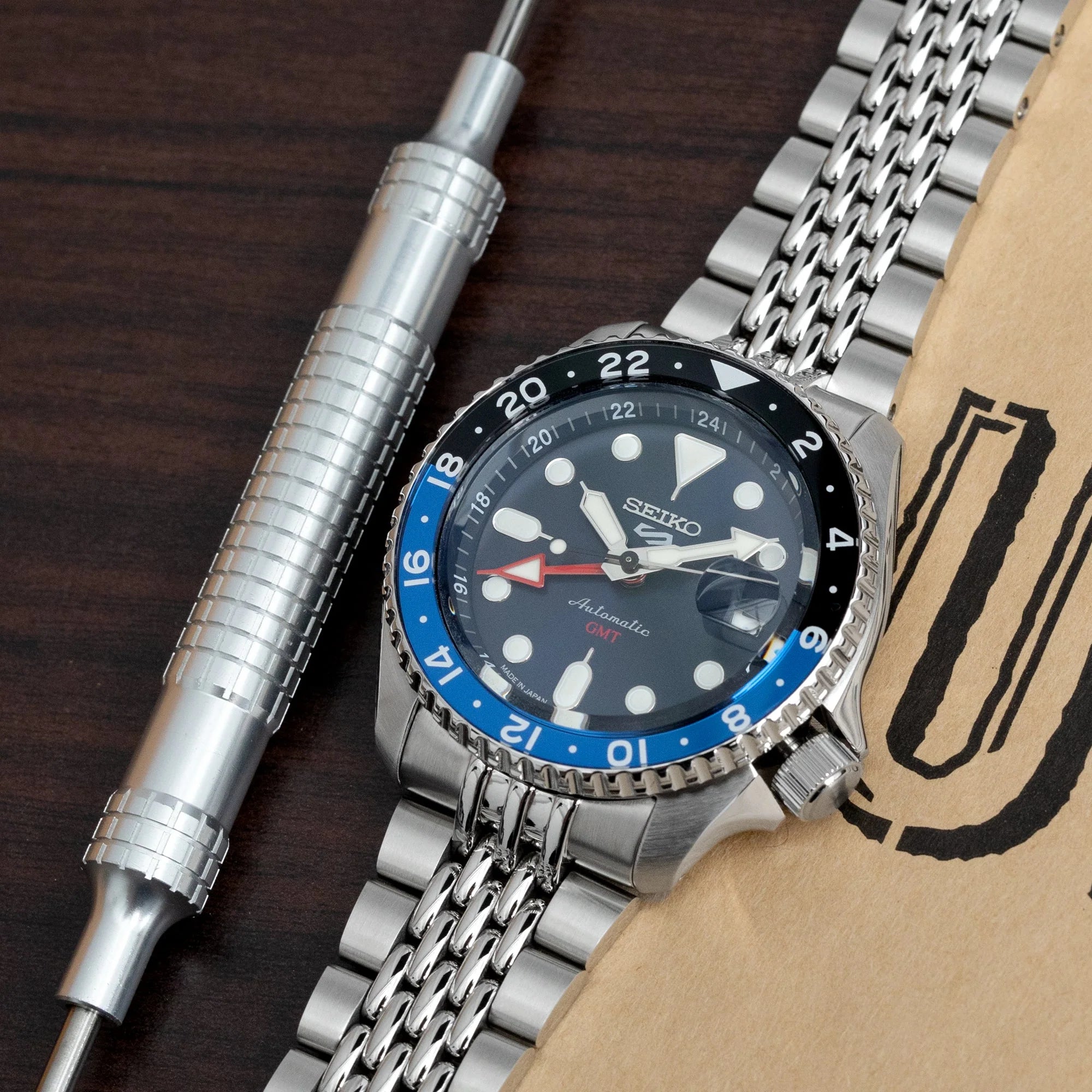 Seiko 5 GMT SSK001 automatic watch with STRAPCODE GOMA BOR steel bracelet and blue black GMT bezel on dark wood background