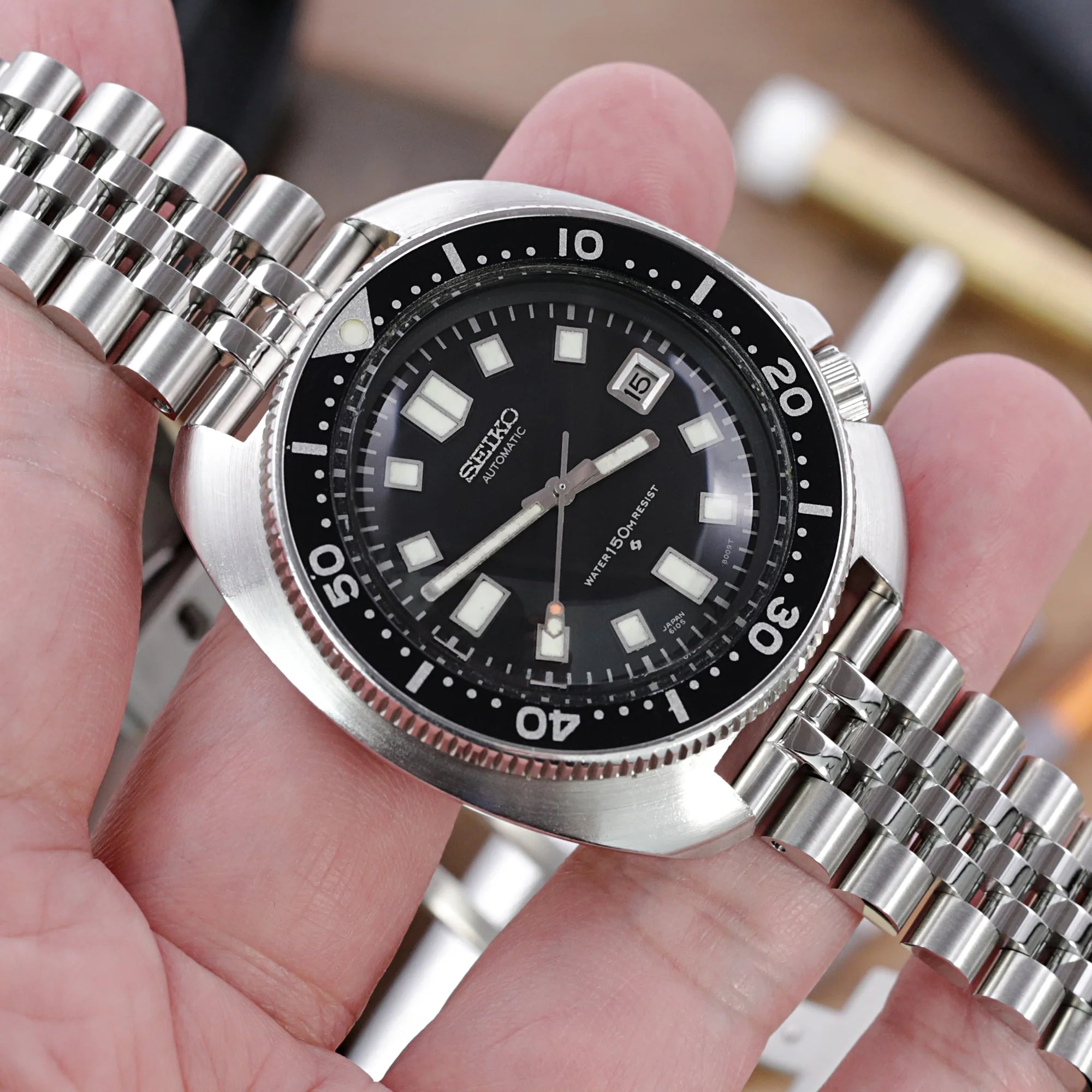Strapcode Super-Jub II stainless steel bracelet with V-clasp fitted on black dial dive watch, showcasing polished cylindrical links and secure fastening mechanism