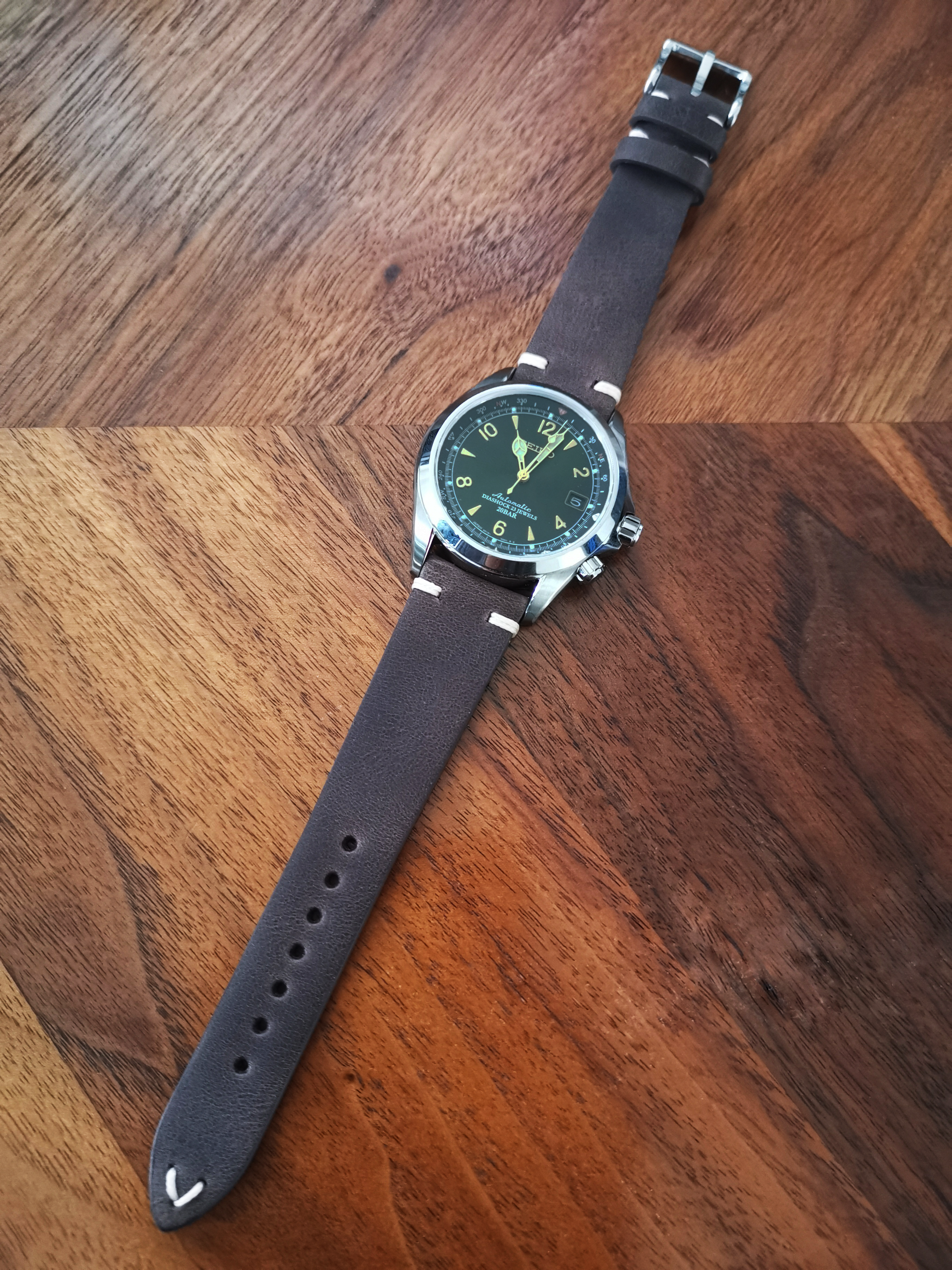 Vintage military-style watch with dark green dial, silver case, and dark brown leather quick release strap on wooden background