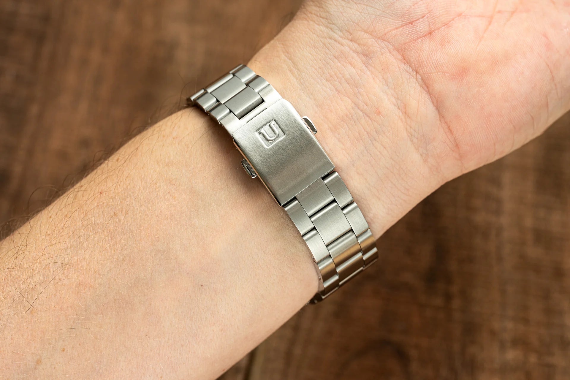 Stainless steel watch bracelet with push-button clasp worn on wrist, featuring brushed metal links and logo detail