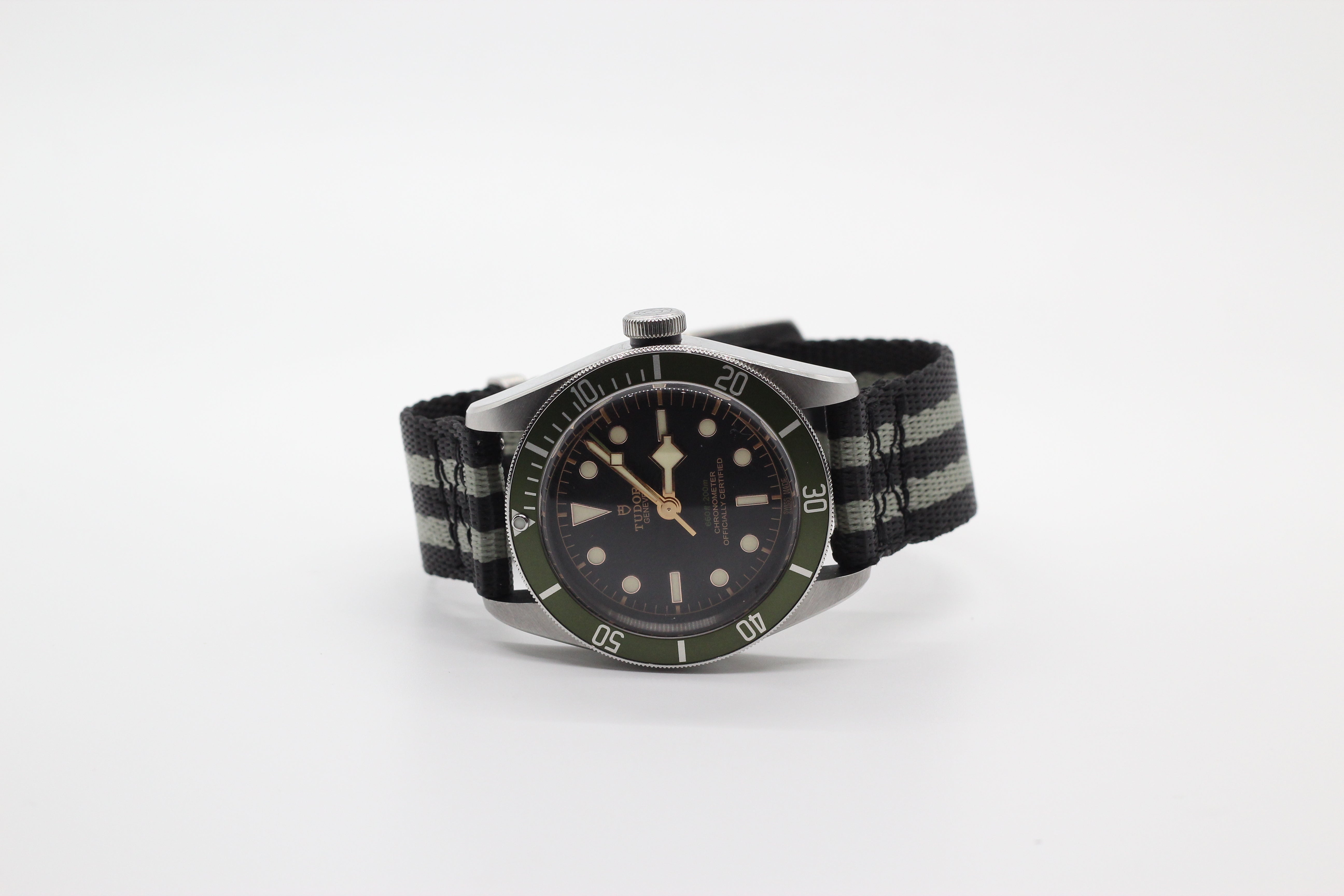 Vintage-style dive watch with black dial, green bezel, and black striped military fabric strap on white background