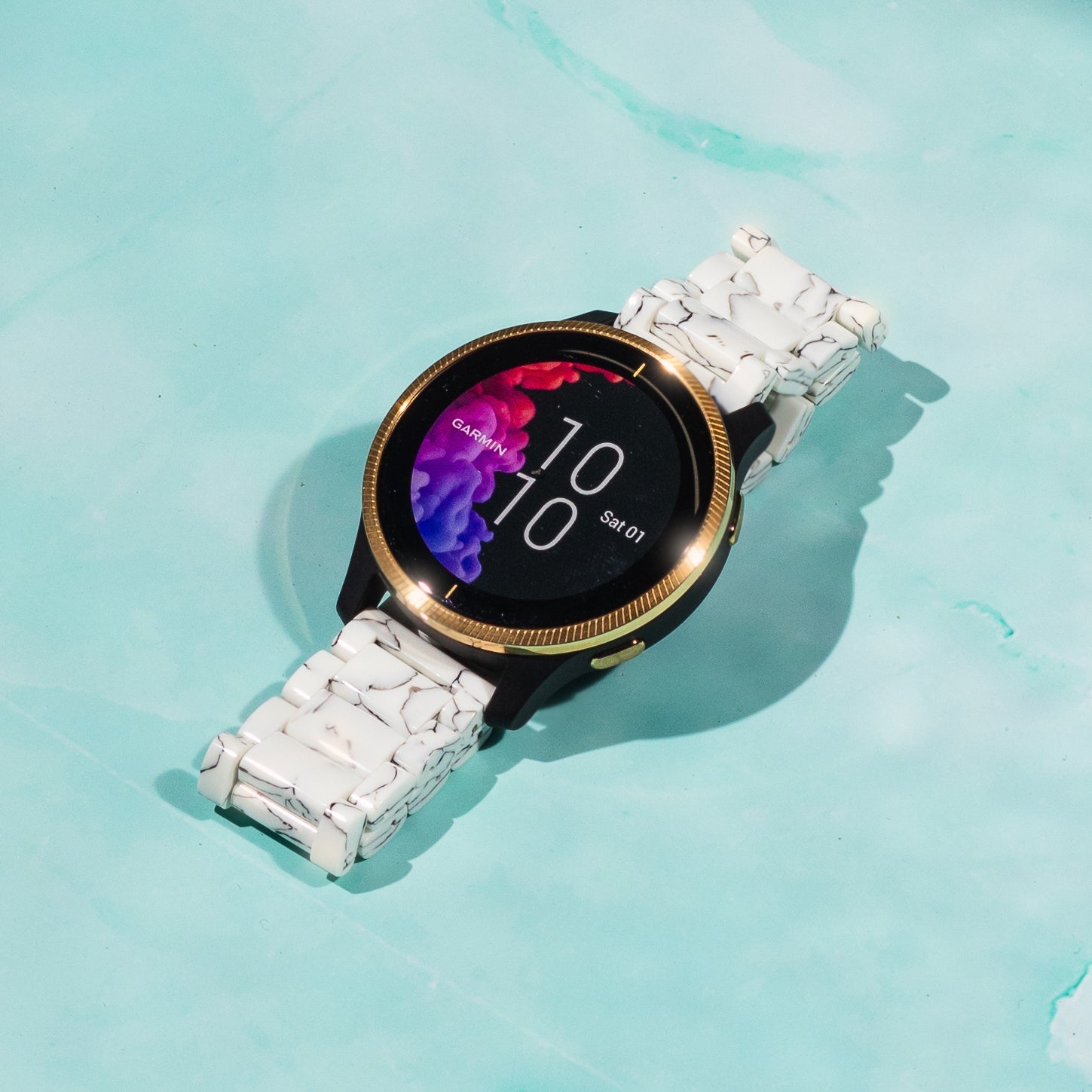Garmin smartwatch with gold bezel and white tortoiseshell acetate band displaying time on turquoise background