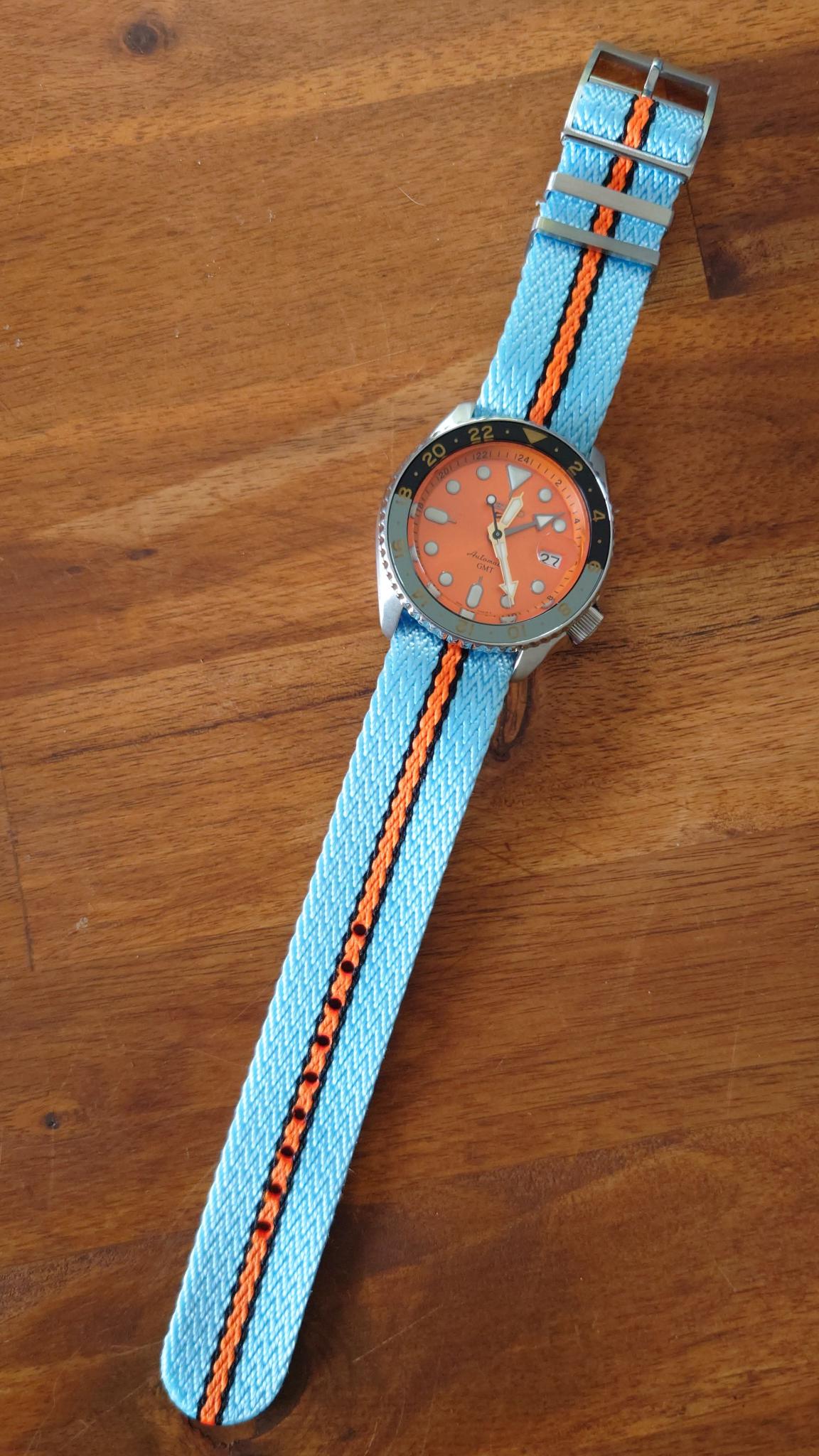 Sports watch with orange dial, stainless steel case, and sky blue fabric strap with orange stripe on wooden background